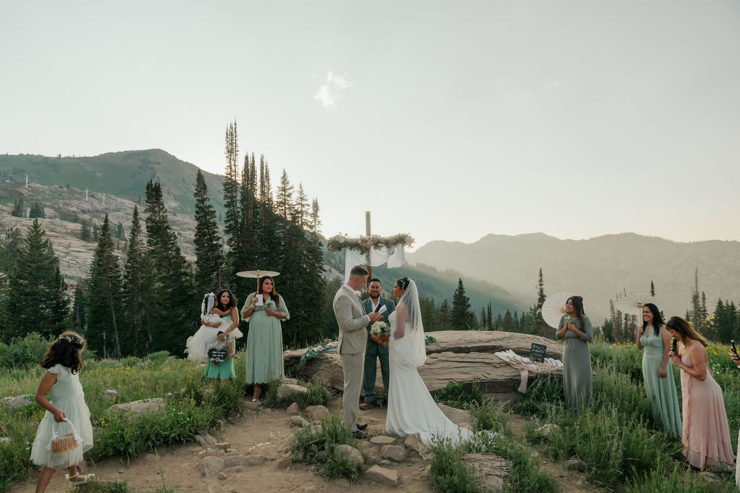 Outdoor mountain wedding ceremony with bridal party in mint dresses surrounded by pine trees and scenic peaks at sunset.