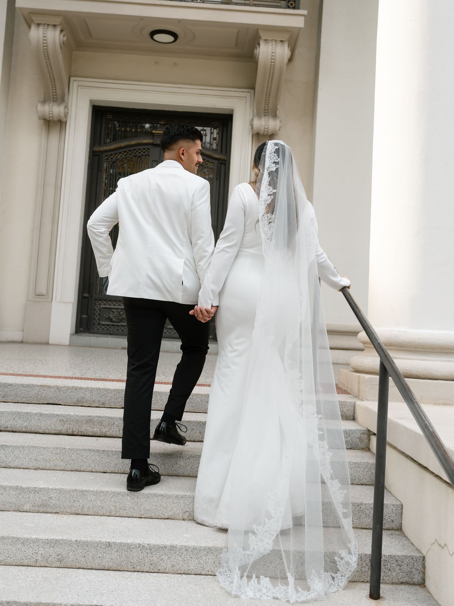 A bride and groom walk hand-in-hand up marble steps toward an elegant building entrance on their wedding day.
