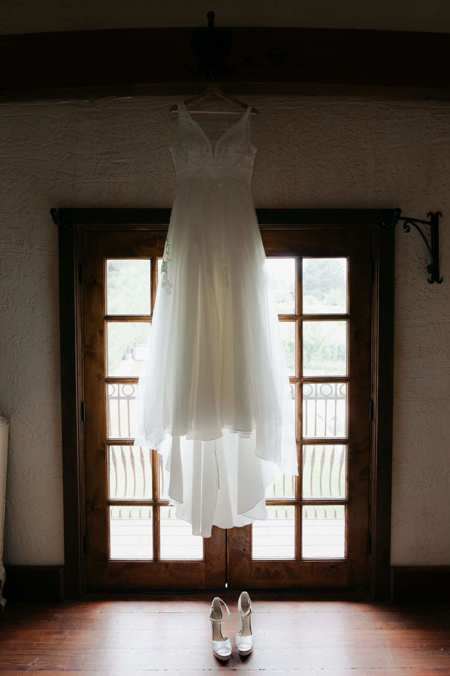 White wedding dress hanging by a window above white bridal heels on a hardwood floor.