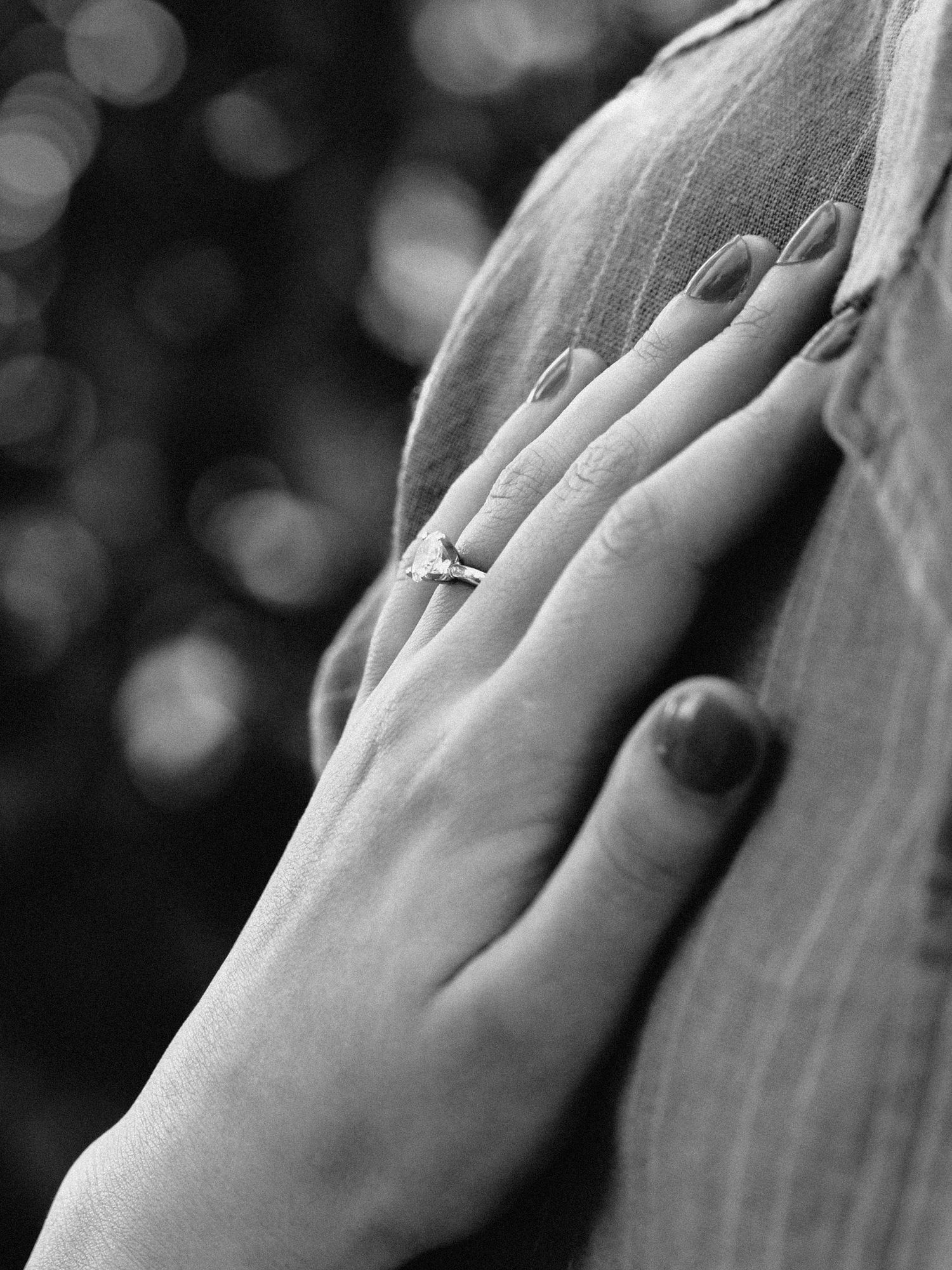 Black and white close-up of a woman's hand with dark nails wearing a diamond engagement ring resting on a shirt.