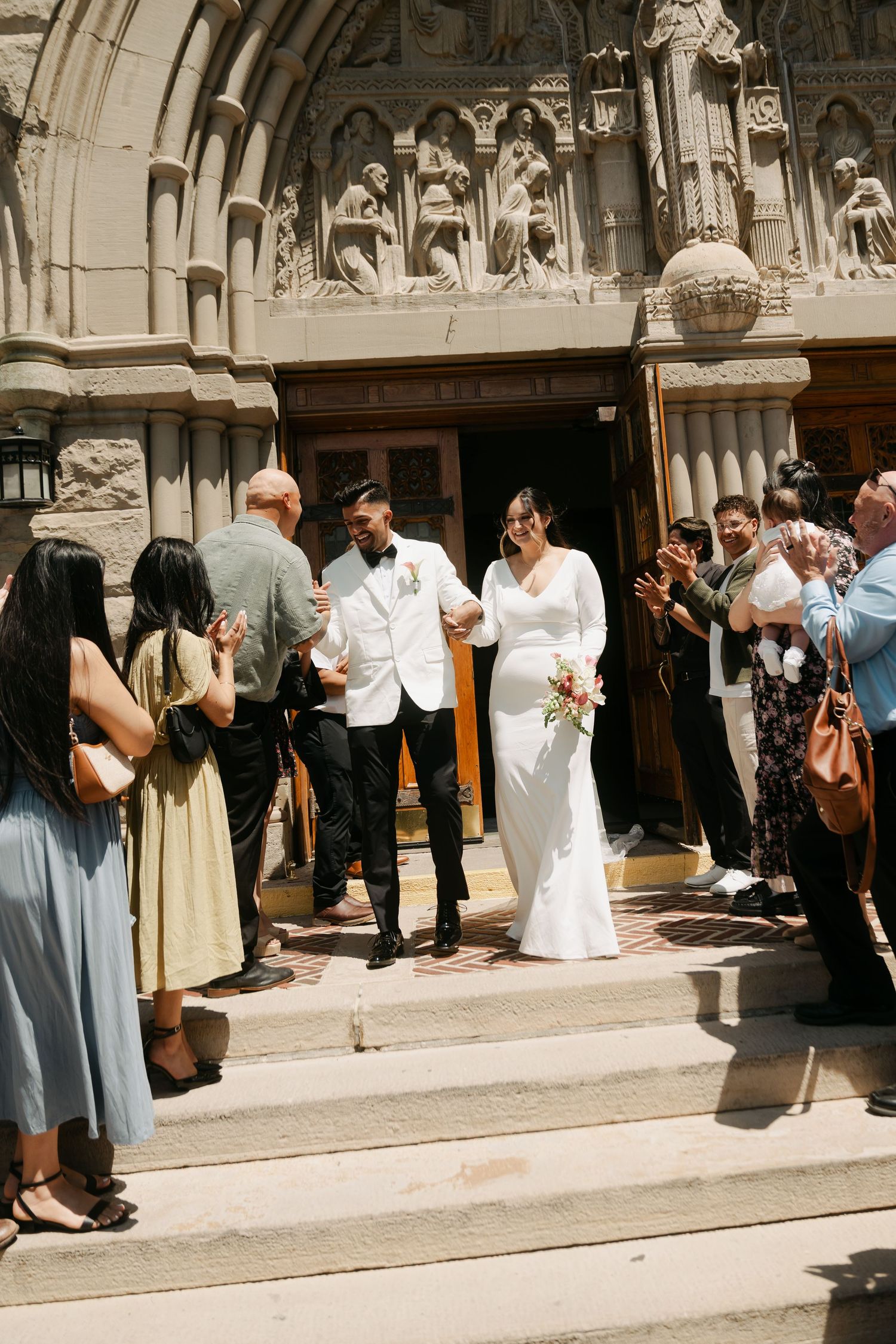 Bride and groom exit ornate church doors, greeted by cheering guests on sunny wedding day.