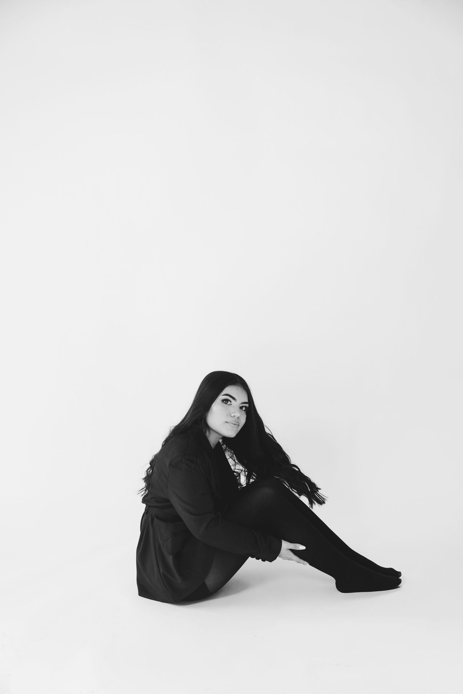 Black and white portrait of a young woman with long dark hair sitting on the floor in a black outfit.