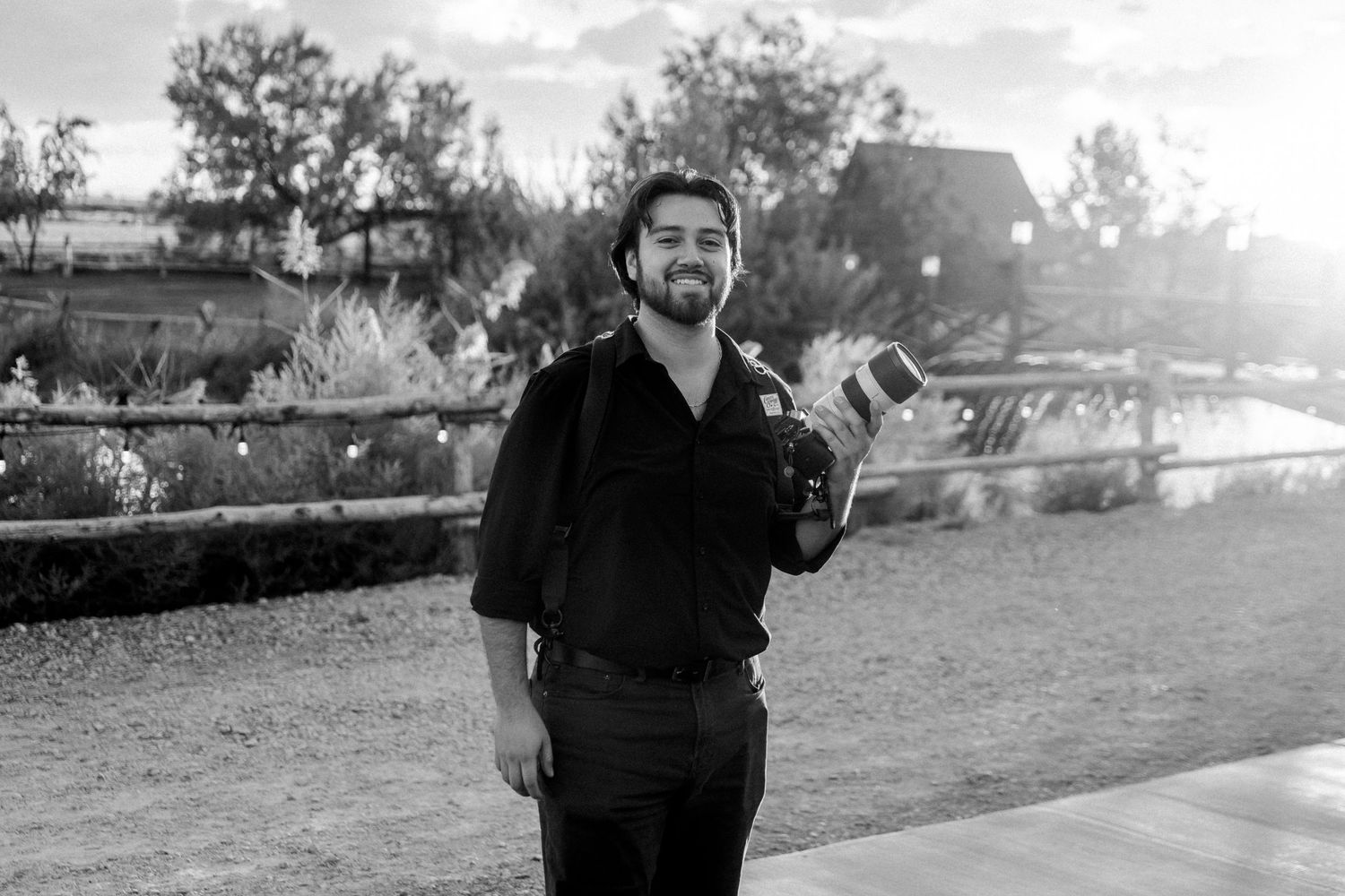 Smiling man holding a camera outdoors in a black and white photo at a rustic venue.