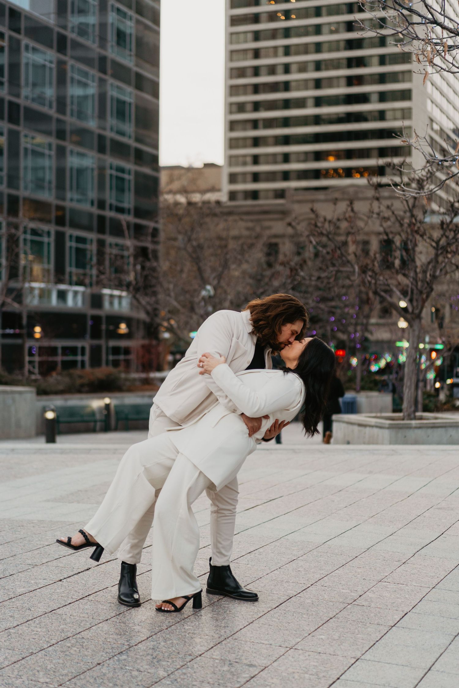 Same-sex couple sharing a romantic dip kiss in an urban plaza with city buildings backdrop.