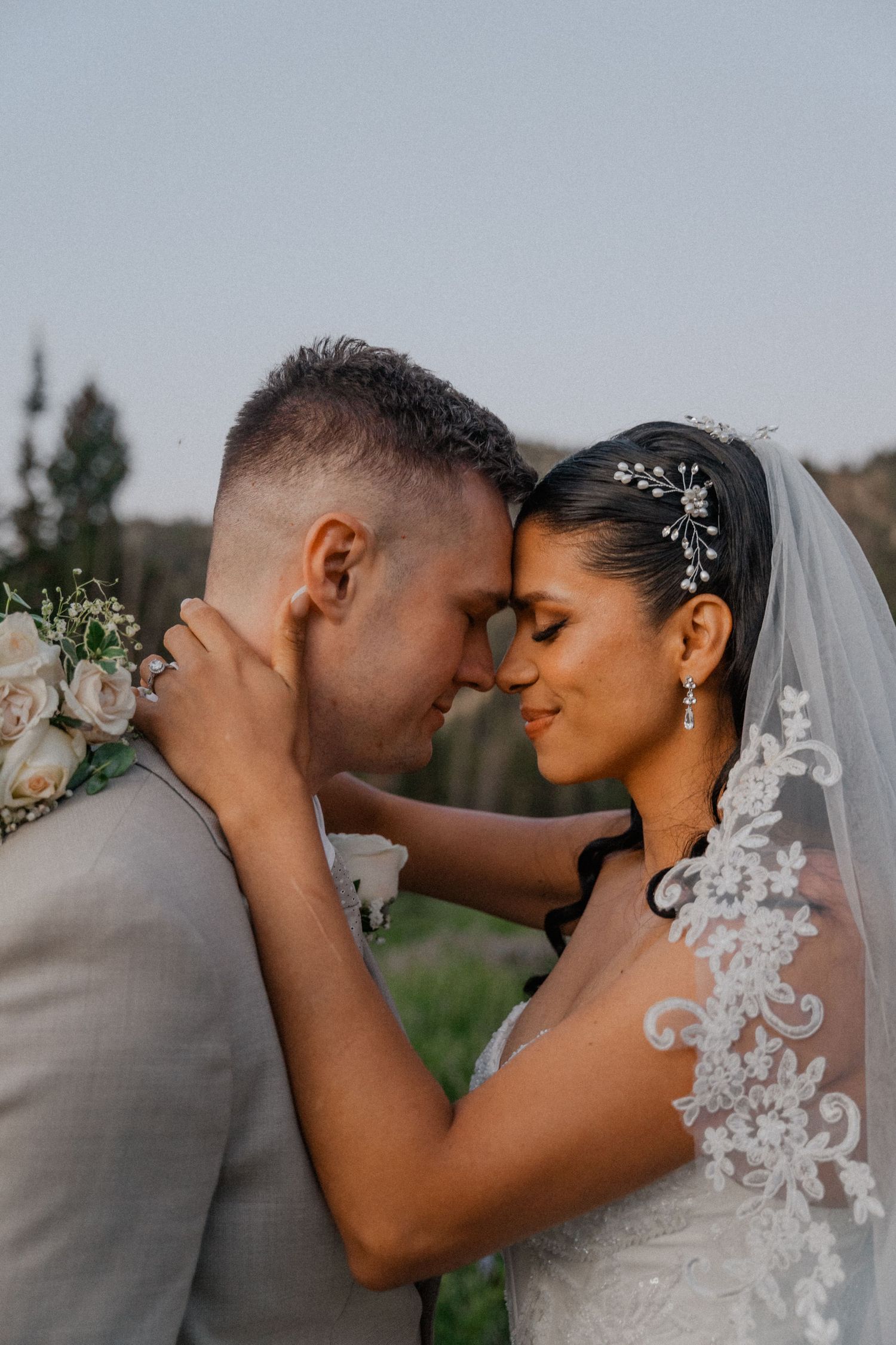 Bride and groom touching foreheads tenderly outdoors, bride wearing lace veil and floral hair piece on wedding day.
