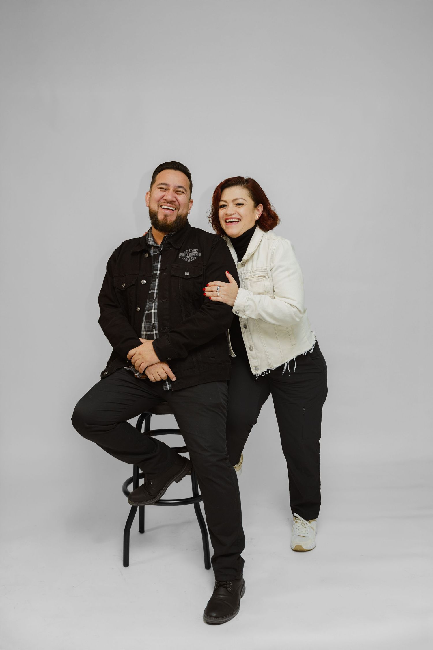 A laughing couple poses together in a studio, dressed in black and white casual jackets.