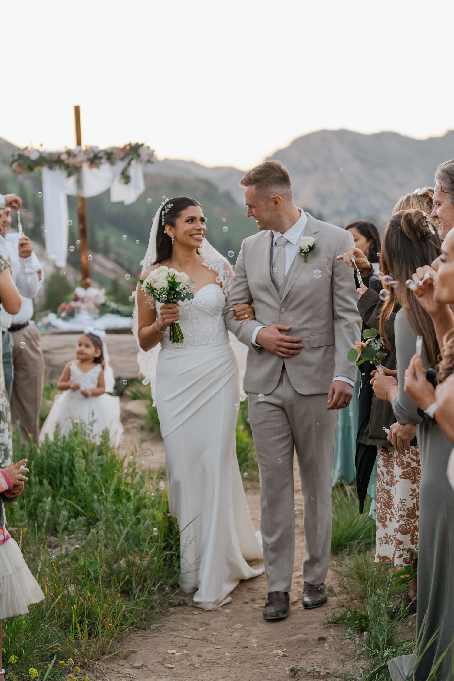 Bride and groom walk down outdoor aisle after wedding ceremony with mountain backdrop and guests celebrating.