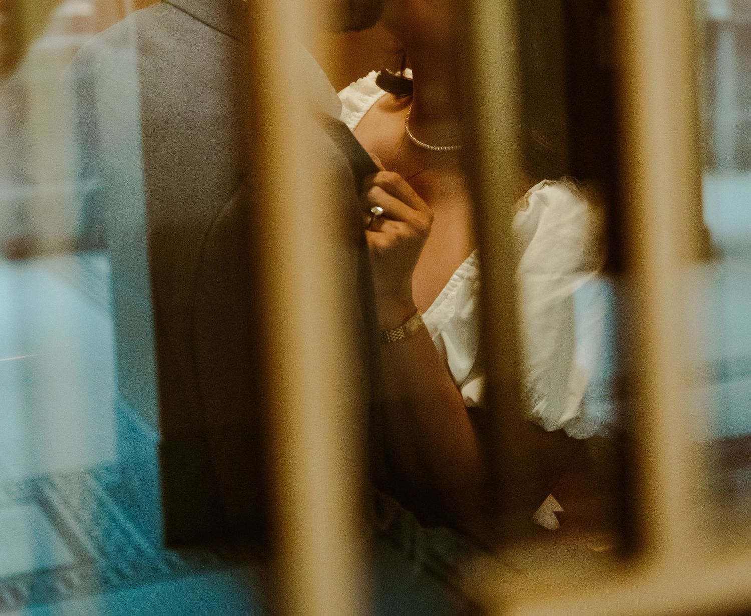 A woman in a white off-shoulder top holds a phone, viewed through blurred golden bars in soft light.