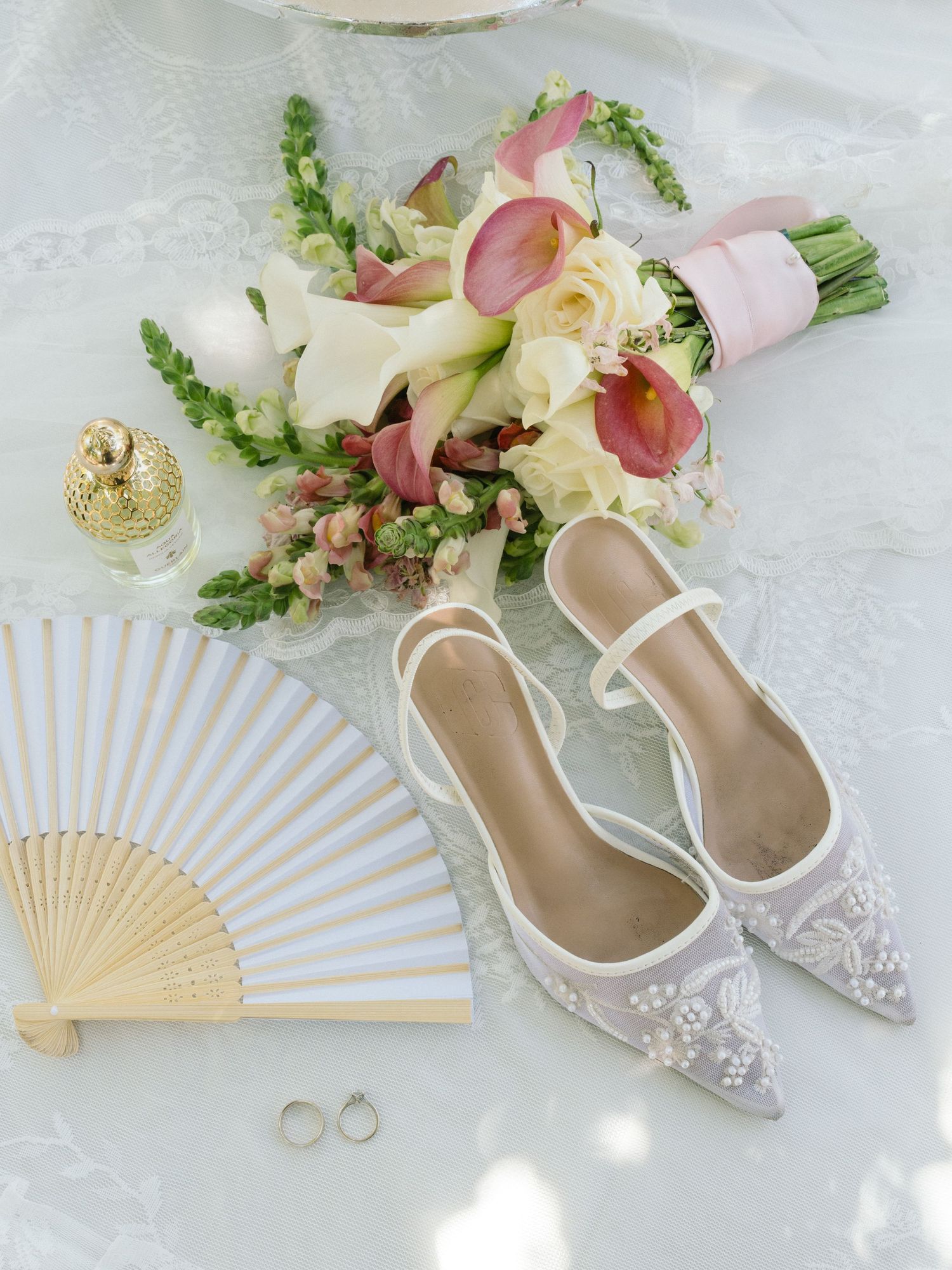 Elegant bridal flat shoes with pearl details beside a pink floral bouquet, hand fan, gold perfume bottle, and wedding rings.