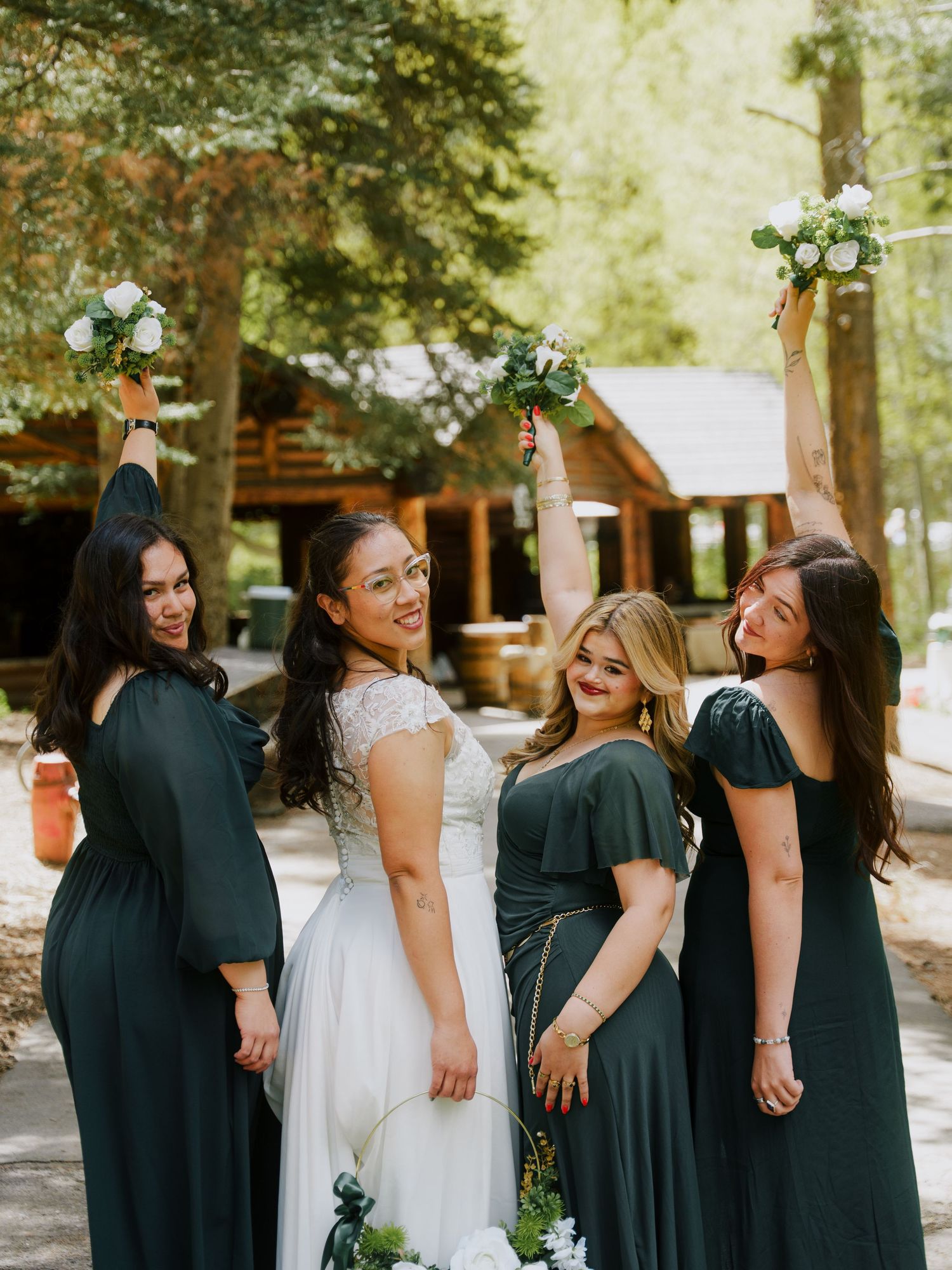 Bride in white lace gown with three bridesmaids in dark green dresses raising bouquets outdoors near a rustic log cabin.