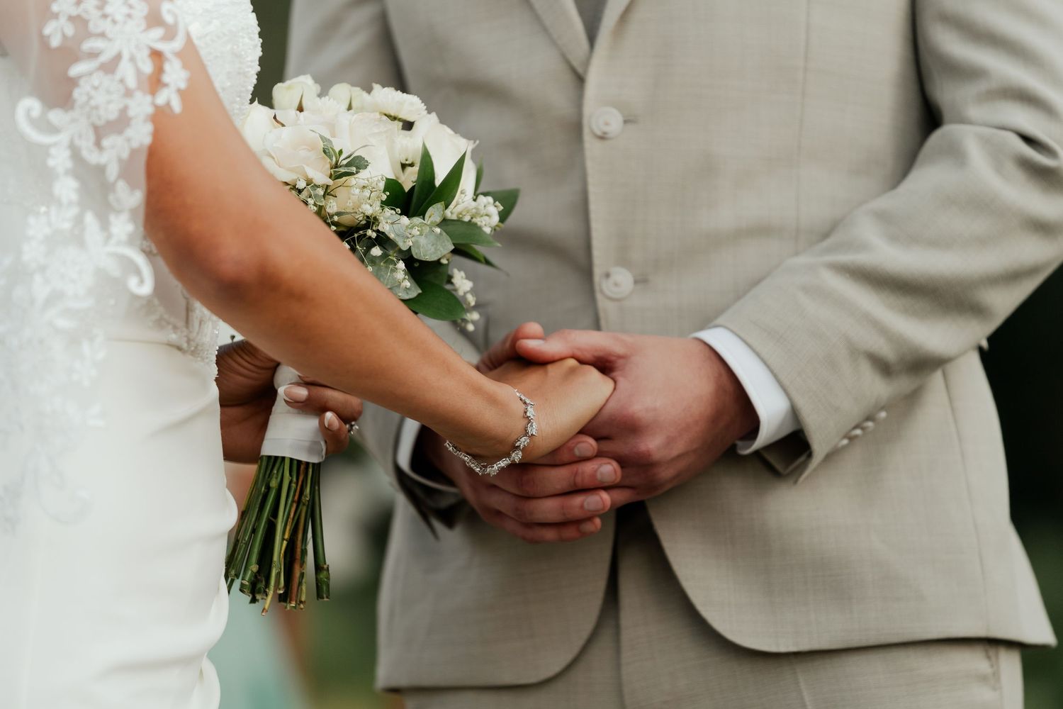 Bride and groom holding hands at wedding ceremony, bride holding white rose bouquet, groom in beige suit.