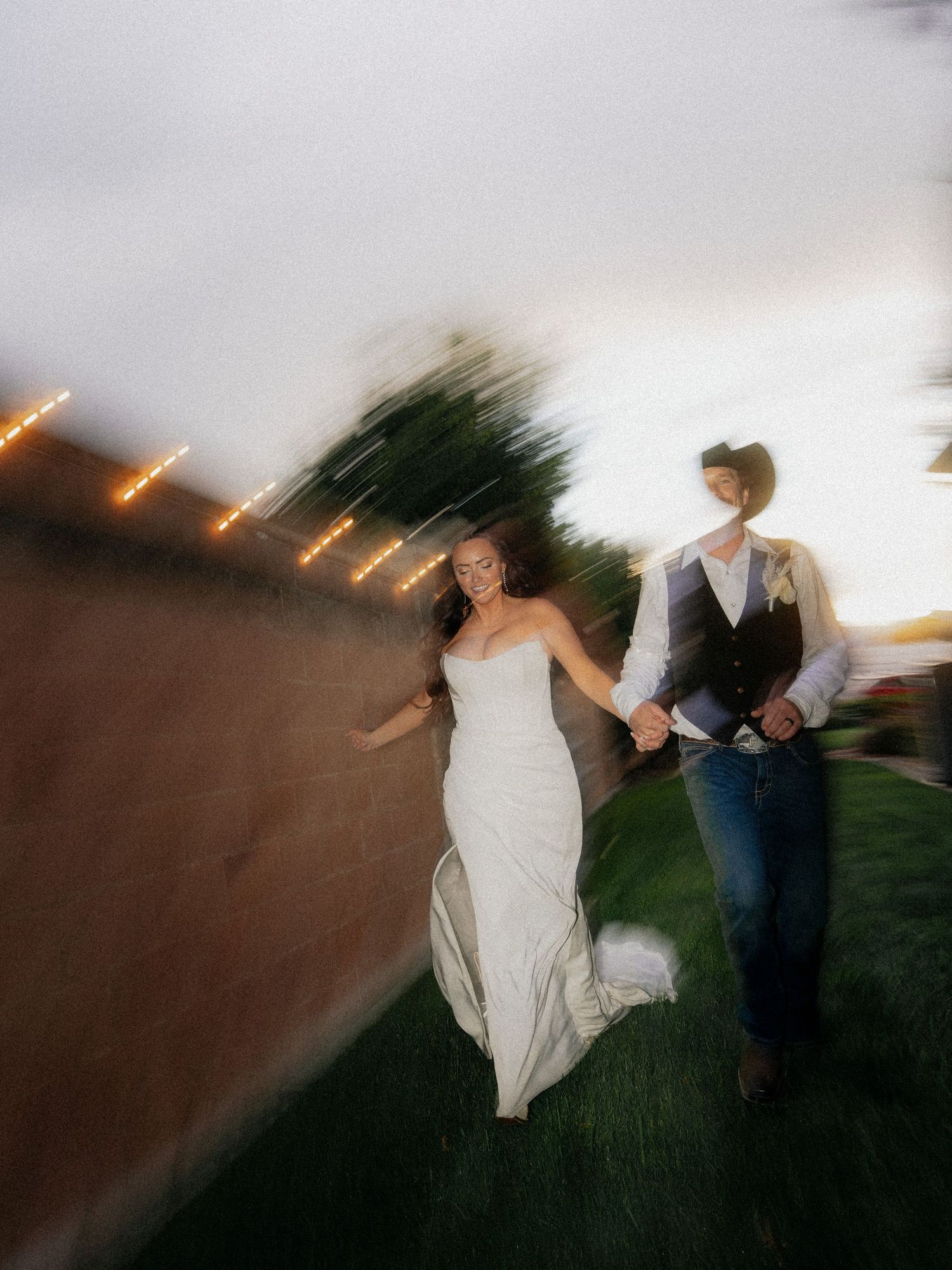 A bride and groom hold hands walking along a rustic wall with string lights at dusk in a motion-blur wedding photo.