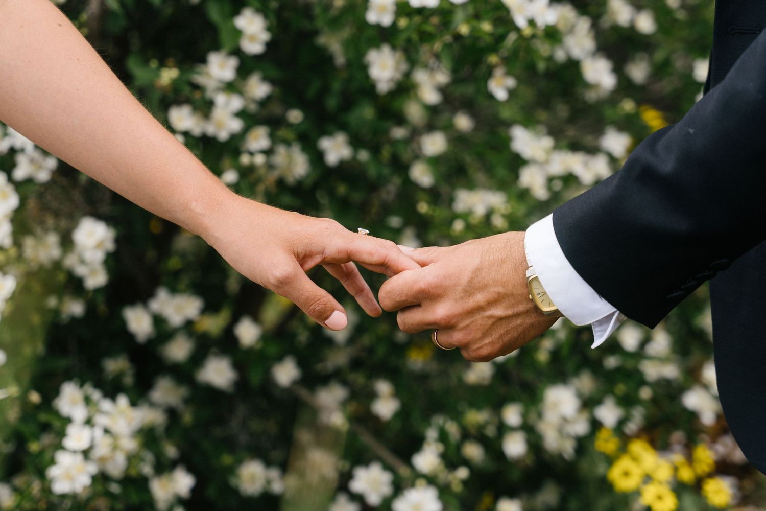 Bride and groom holding hands gently in front of white flowers during outdoor wedding ceremony.