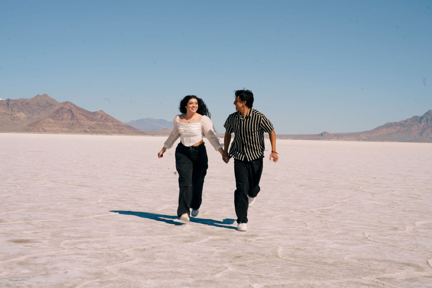 Happy couple holding hands running across vast white salt flats under clear blue sky.