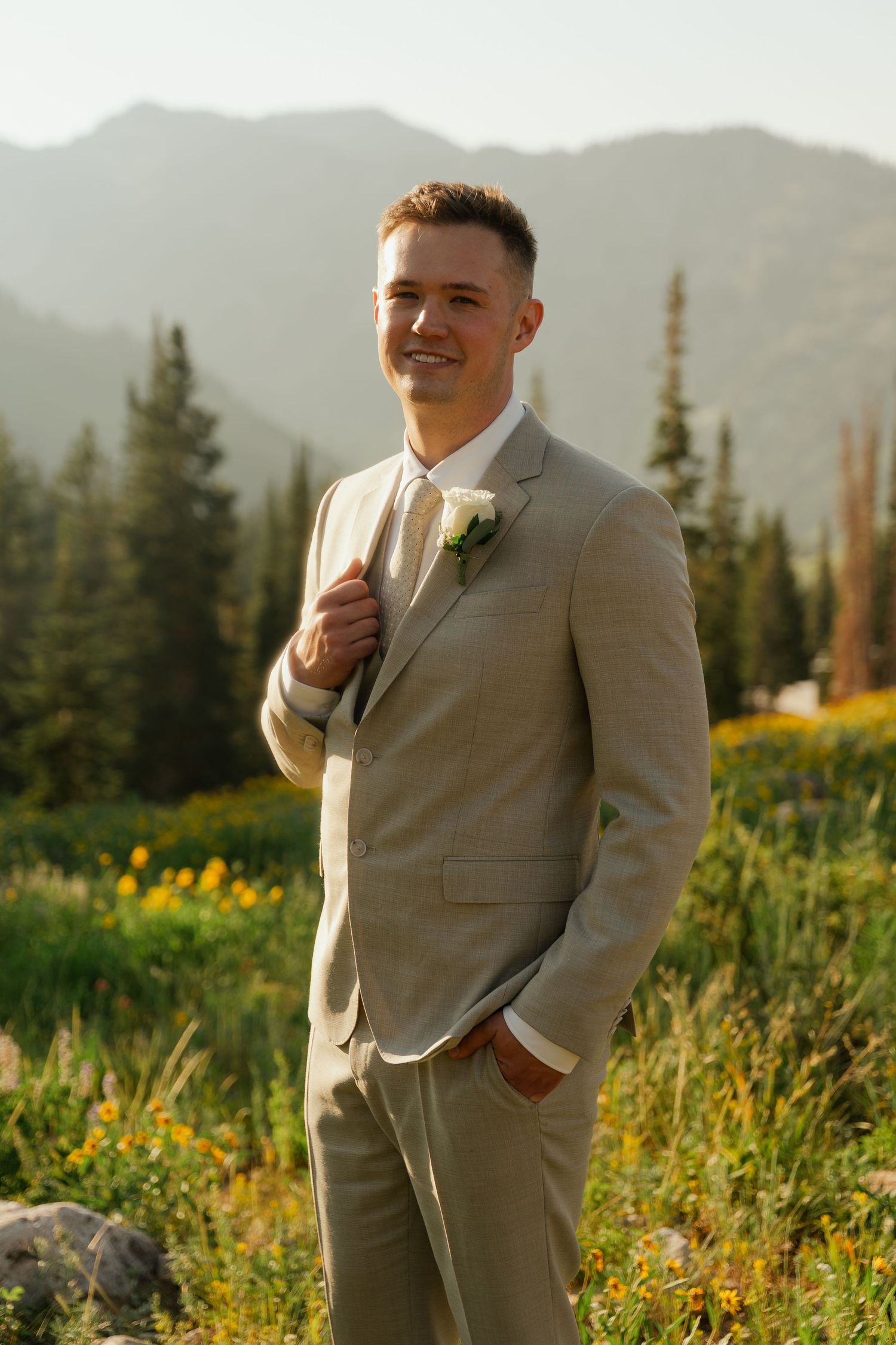 Smiling groom in beige suit with white boutonniere poses outdoors with mountain and wildflower backdrop.