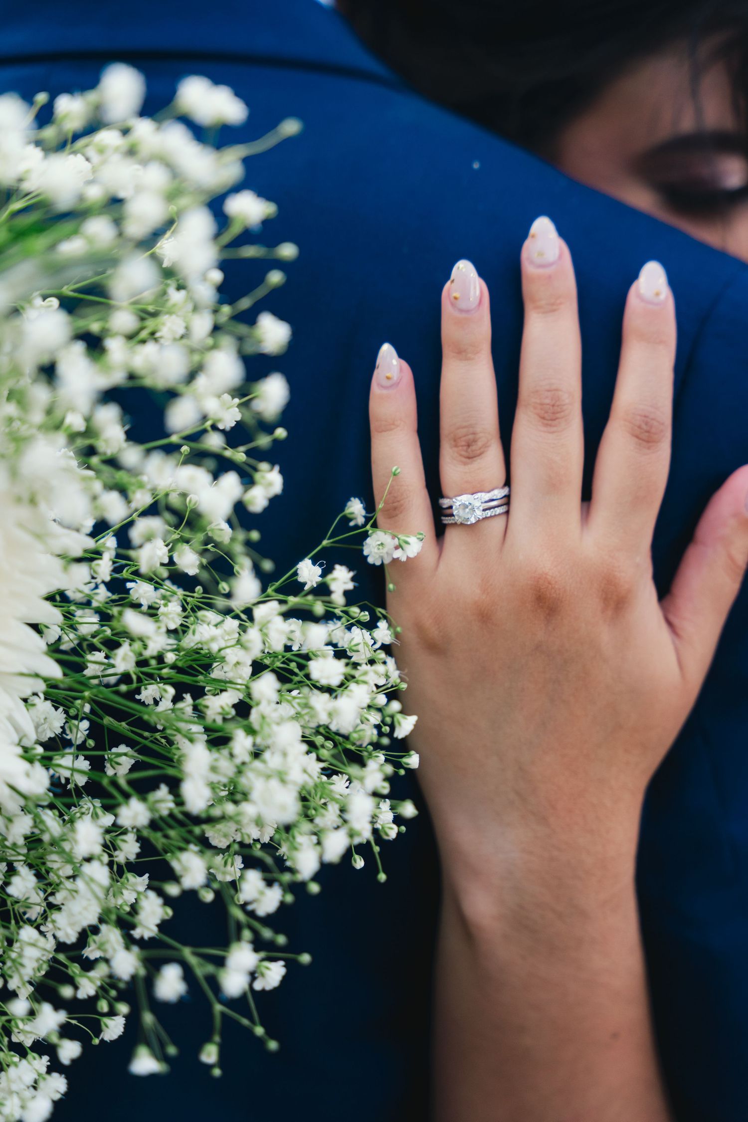 Bride's hand with diamond wedding ring set resting on groom's navy suit beside white baby's breath bouquet.
