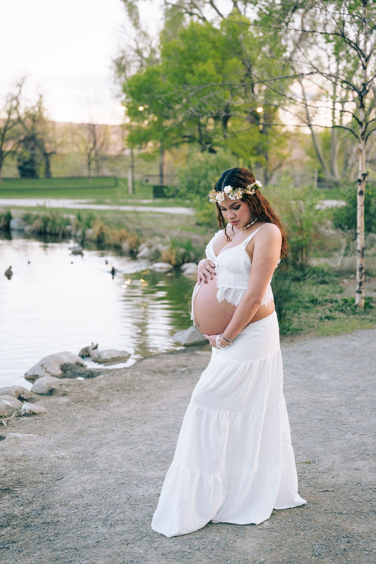 Pregnant woman in white dress and floral crown cradling baby bump beside serene park pond.