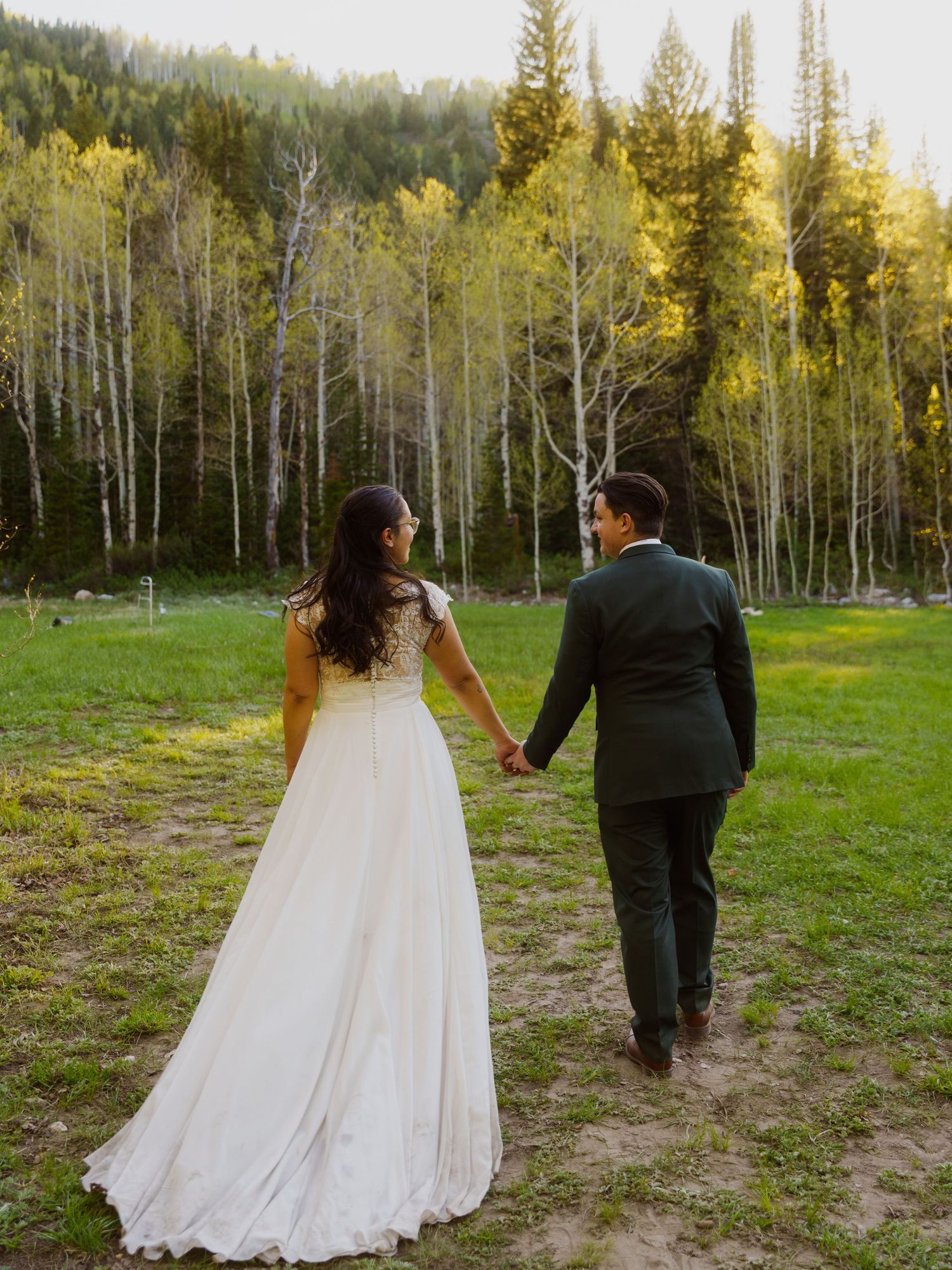 Newlywed couple holding hands walking toward lush green forest in a meadow, bride in white gown and groom in dark suit.