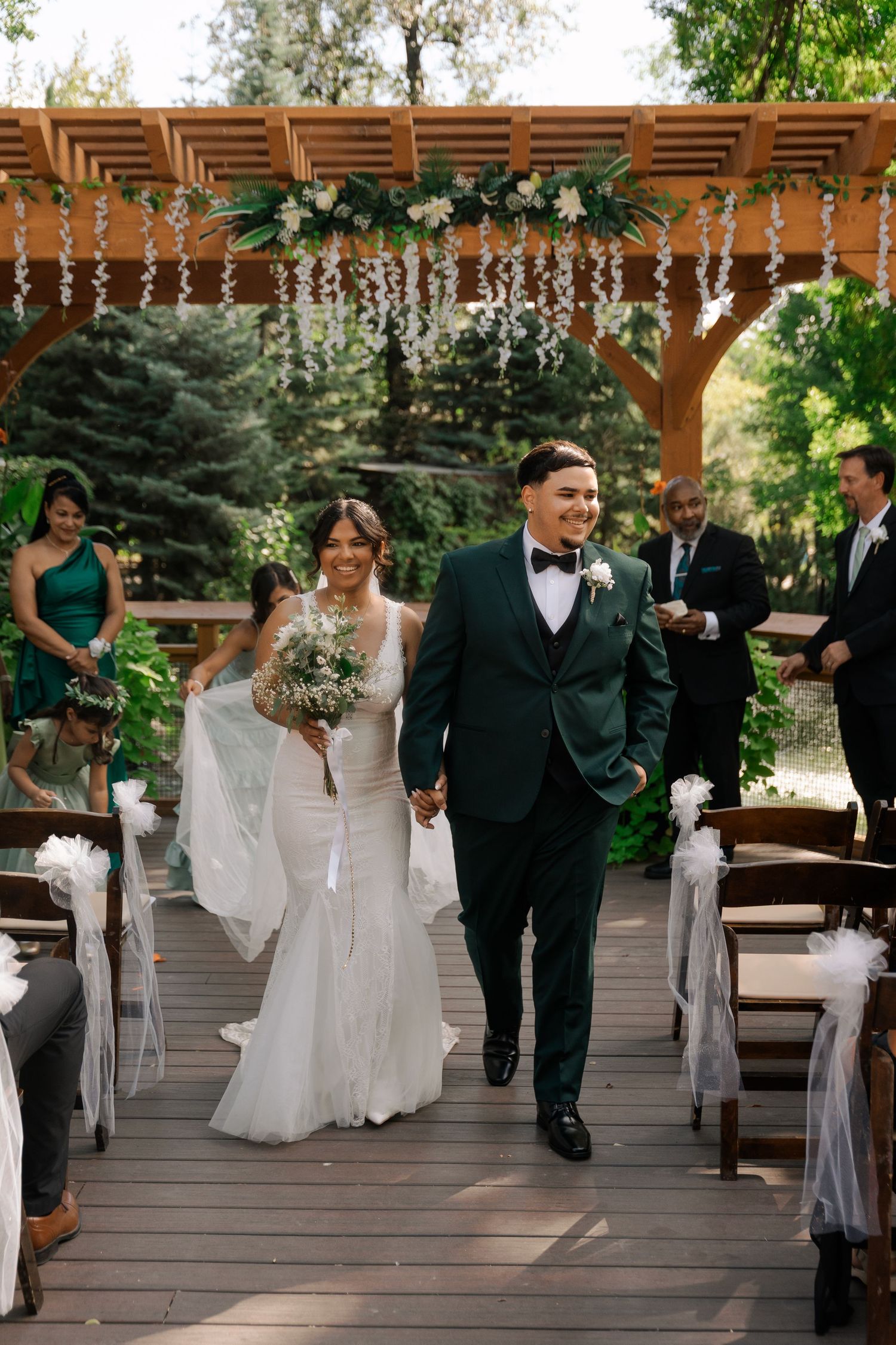 Bride and groom walking down aisle after ceremony under wooden pergola with floral arch.