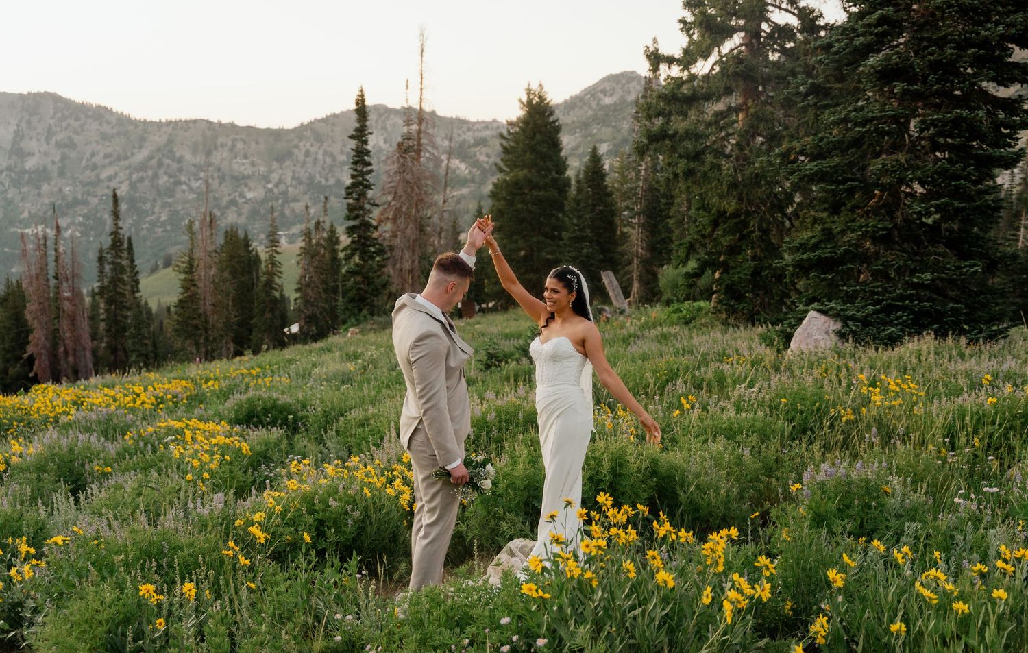 Bride and groom dancing in a mountain wildflower meadow with pine trees at golden hour.