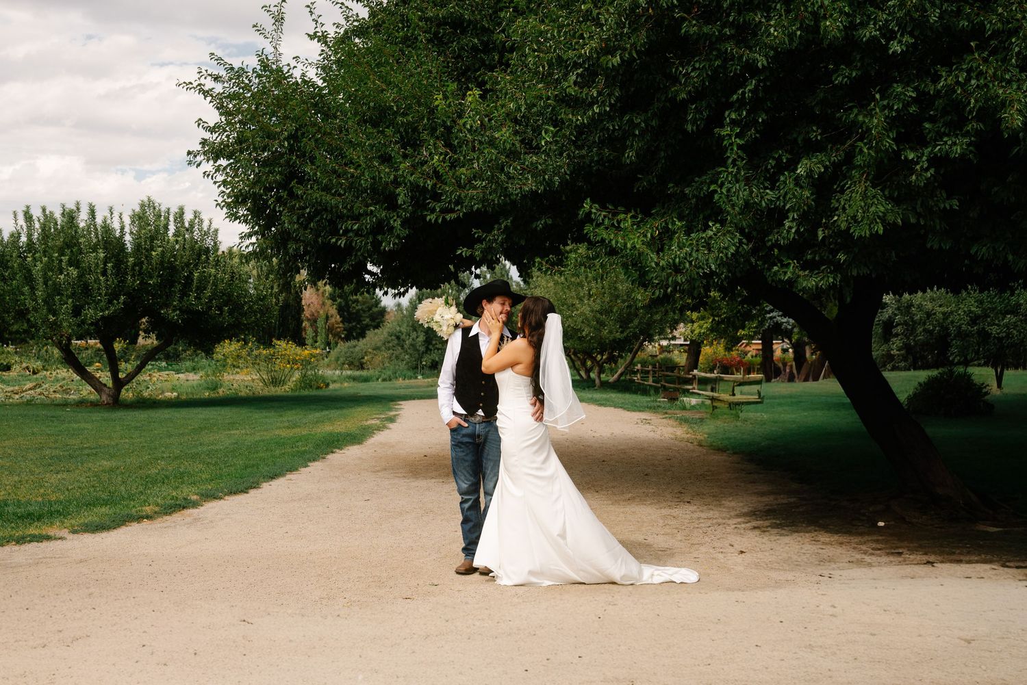 Bride and groom share a romantic kiss on a garden path surrounded by lush green trees on their wedding day.