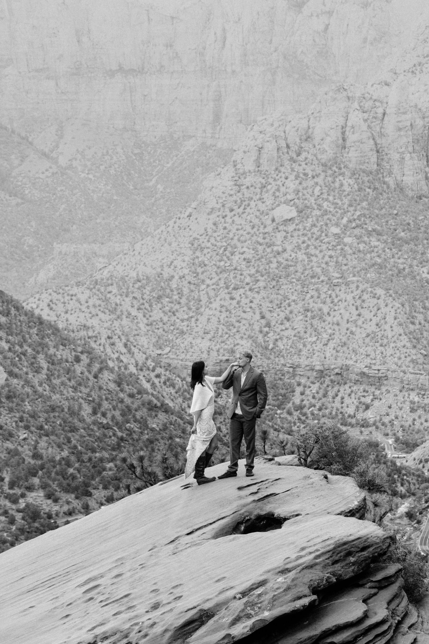 Black and white photo of a couple standing on a rocky cliff edge with a stunning canyon landscape behind them.