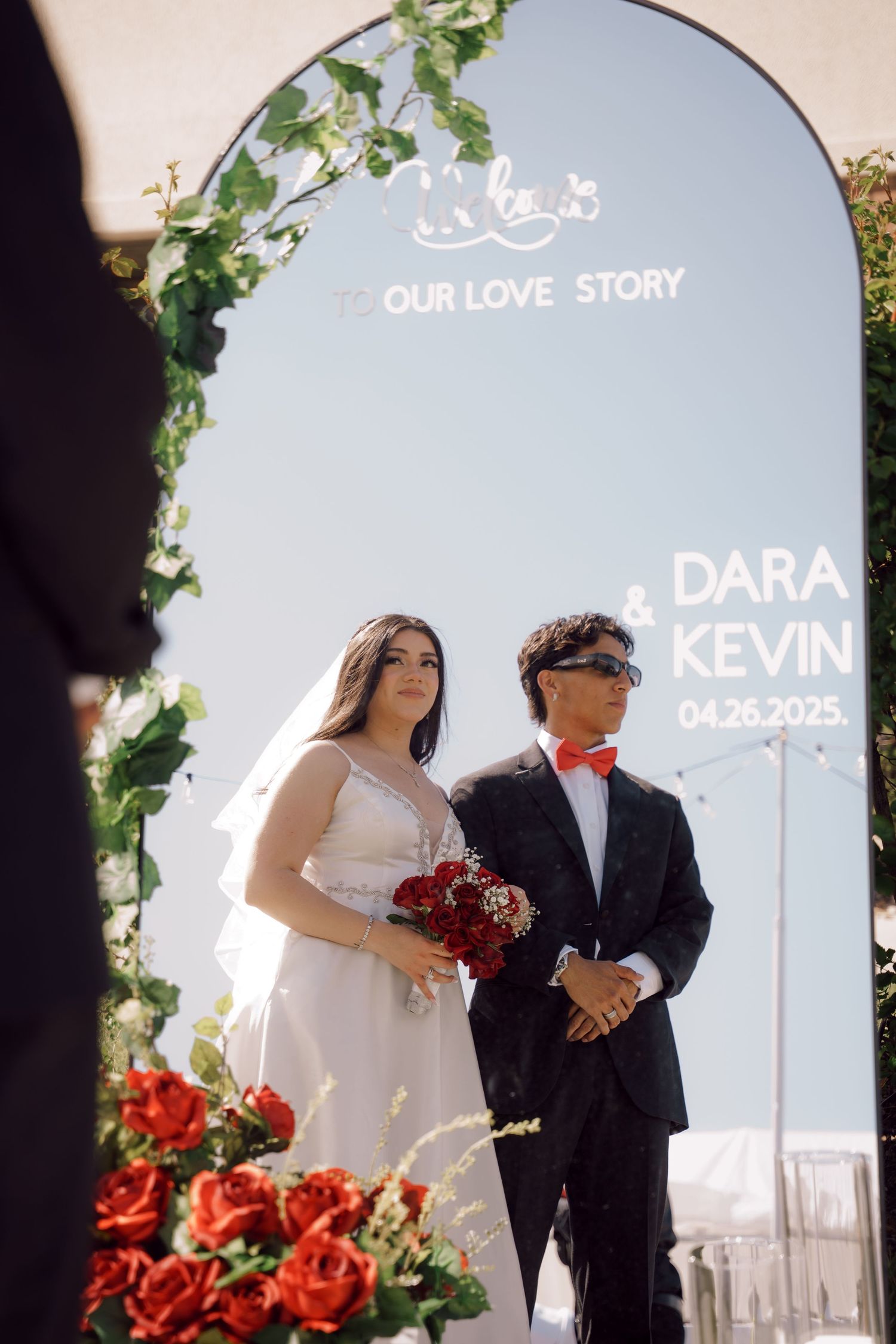 Bride and groom standing under a floral arch with 'Our Love Story' sign at Dara and Kevin's wedding.