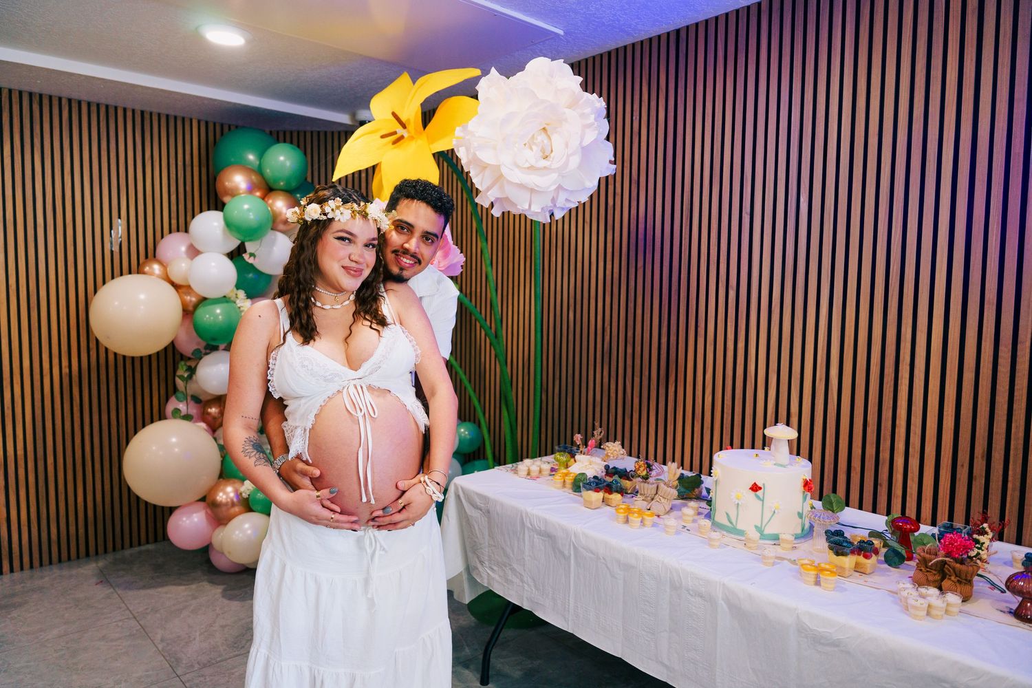 Expecting couple at baby shower embracing bump beside decorated table with balloons and flowers.