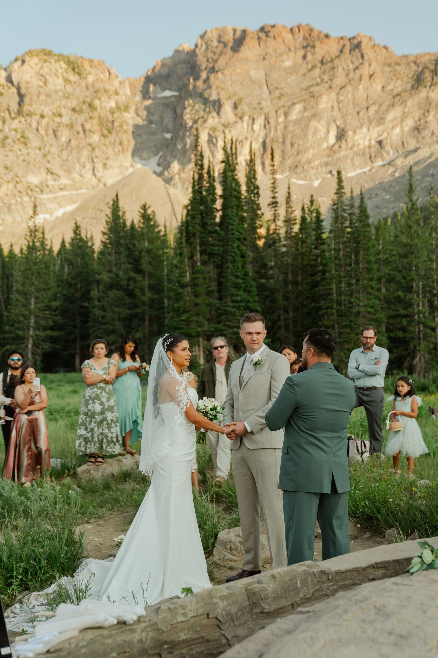 Outdoor mountain wedding ceremony with bride in white gown and veil, groom in tan suit, officiant, and guests amid pine trees.