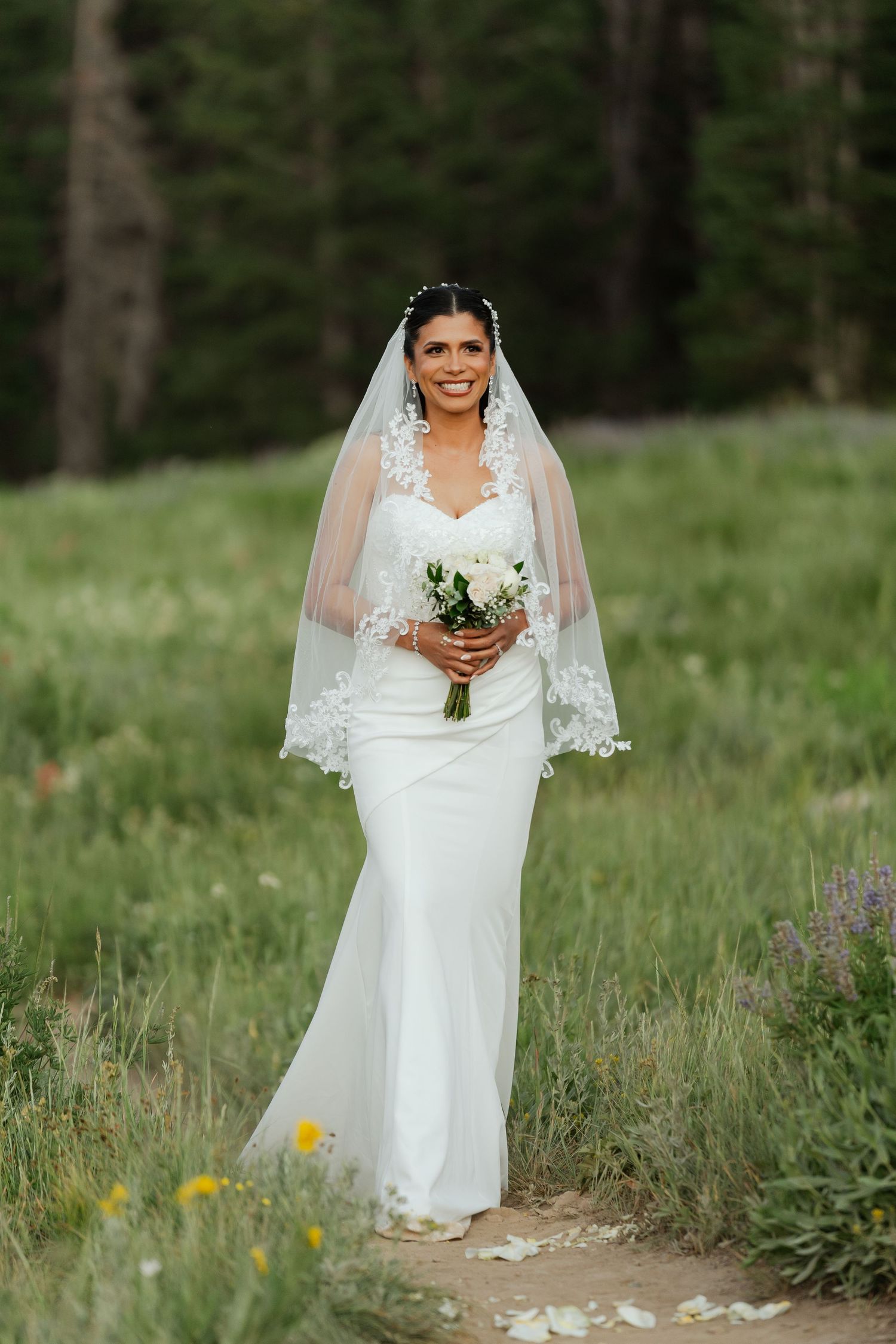 Bride in white lace gown and veil holds bouquet in mountain meadow.