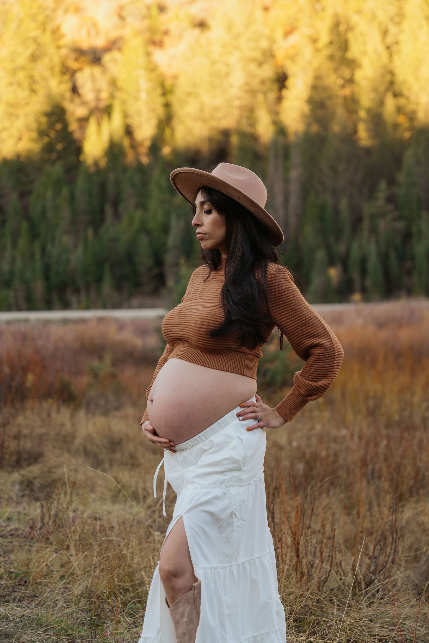 Pregnant woman in brown crop top, white skirt, and wide-brim hat poses outdoors in autumn field with trees.