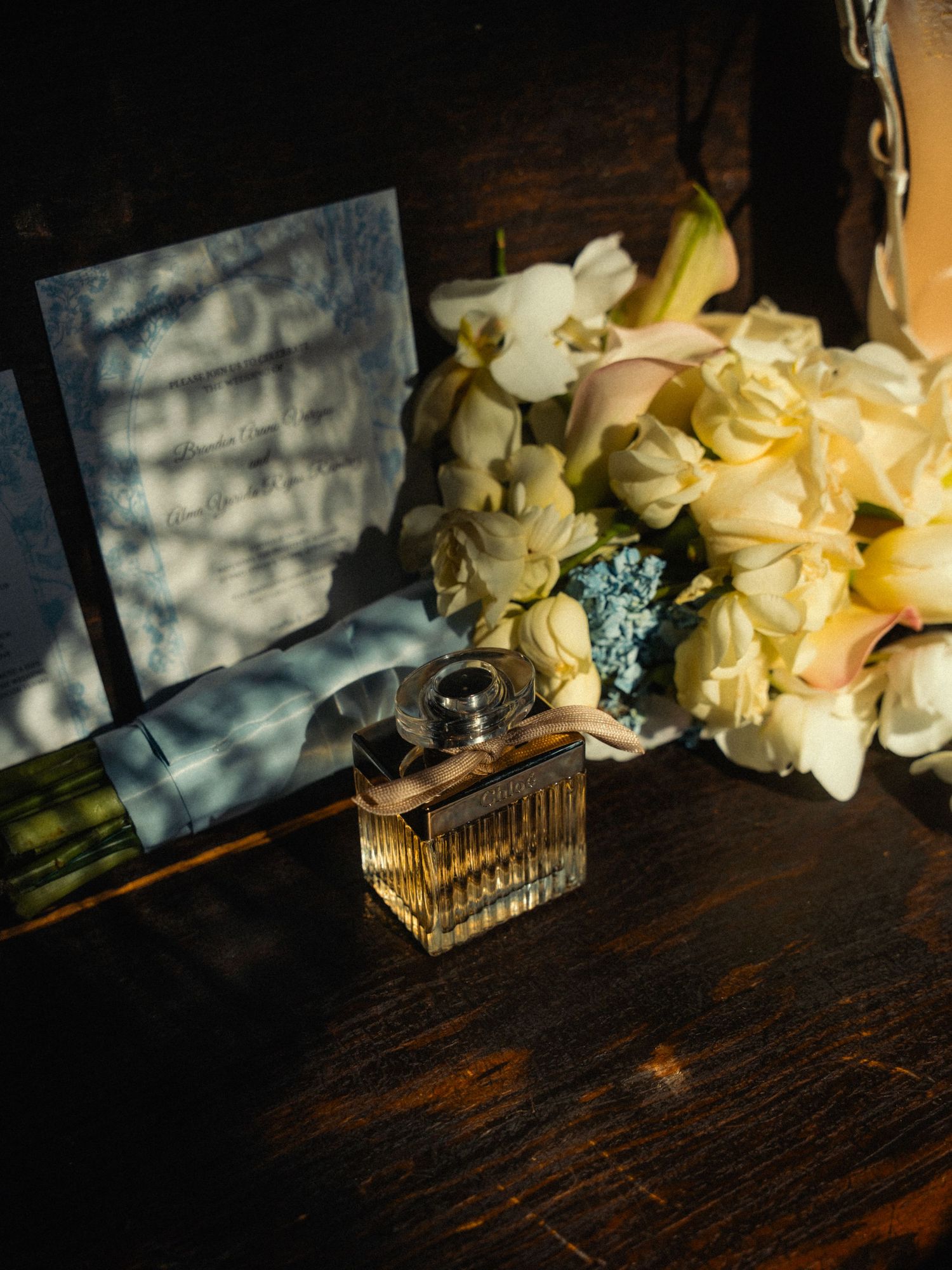 A perfume bottle beside white flowers and a wedding invitation card on a rustic wooden surface in warm light.