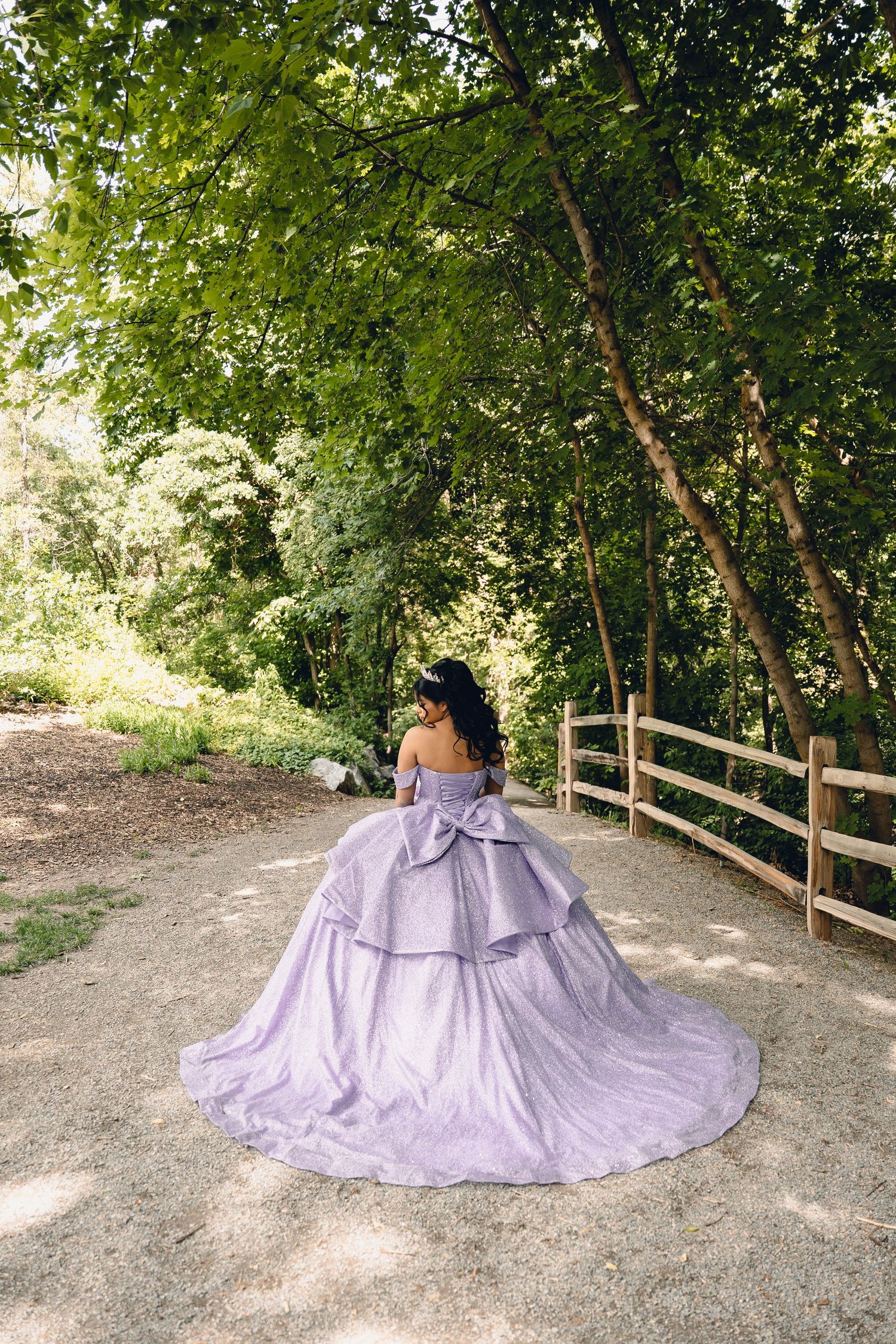 Woman in elegant lavender ball gown with bow standing on a garden path surrounded by green trees.