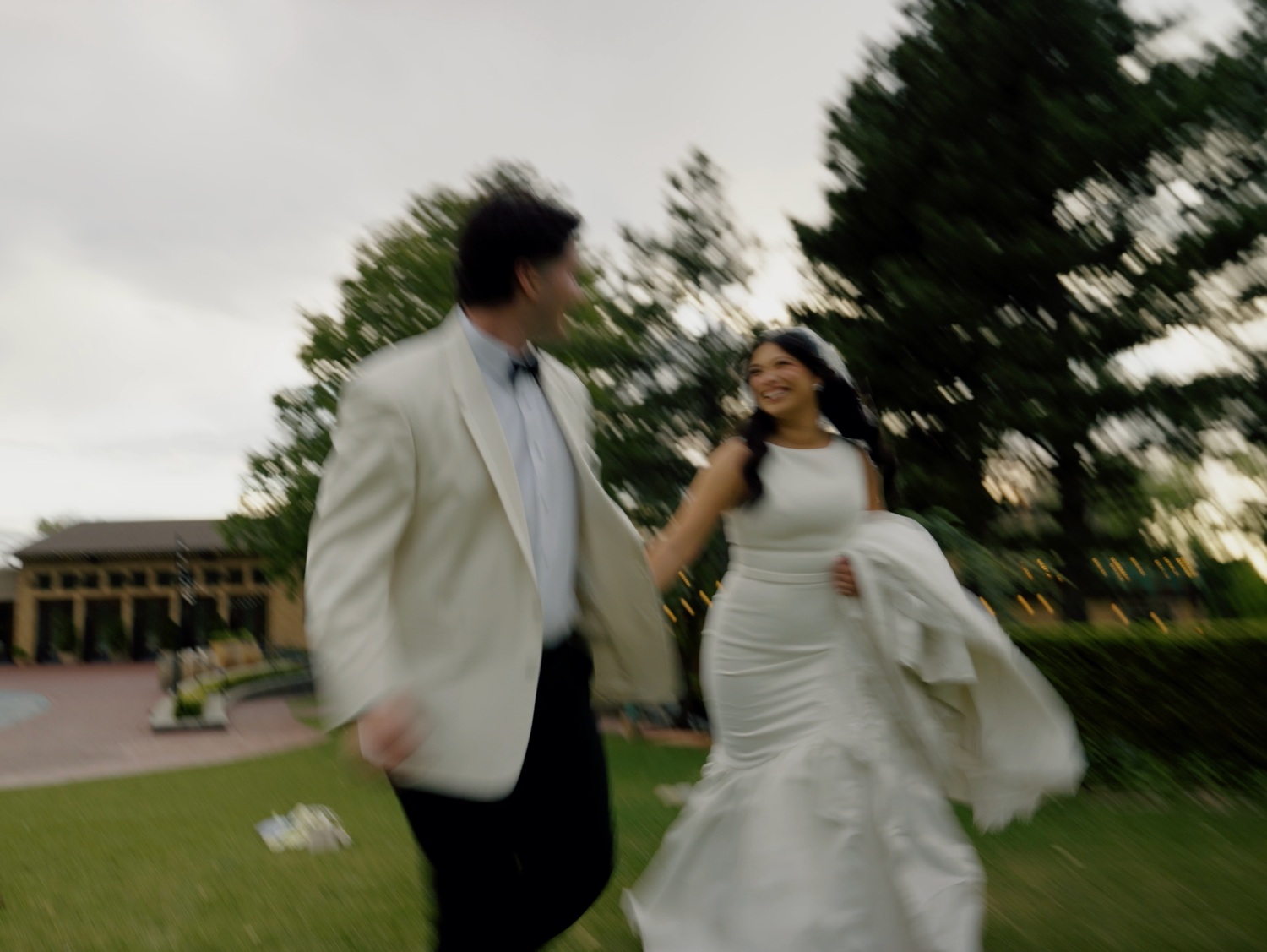 Bride and groom running joyfully on green lawn in wedding attire, motion blur capturing romantic celebration outdoors.