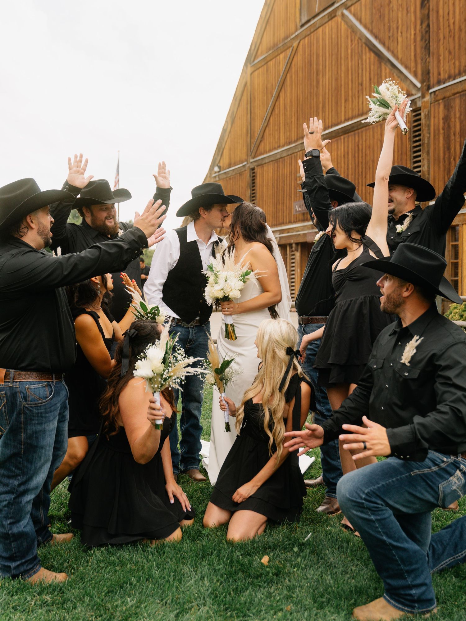 Western wedding couple kissing surrounded by cheering bridal party in black cowboy hats near rustic barn.