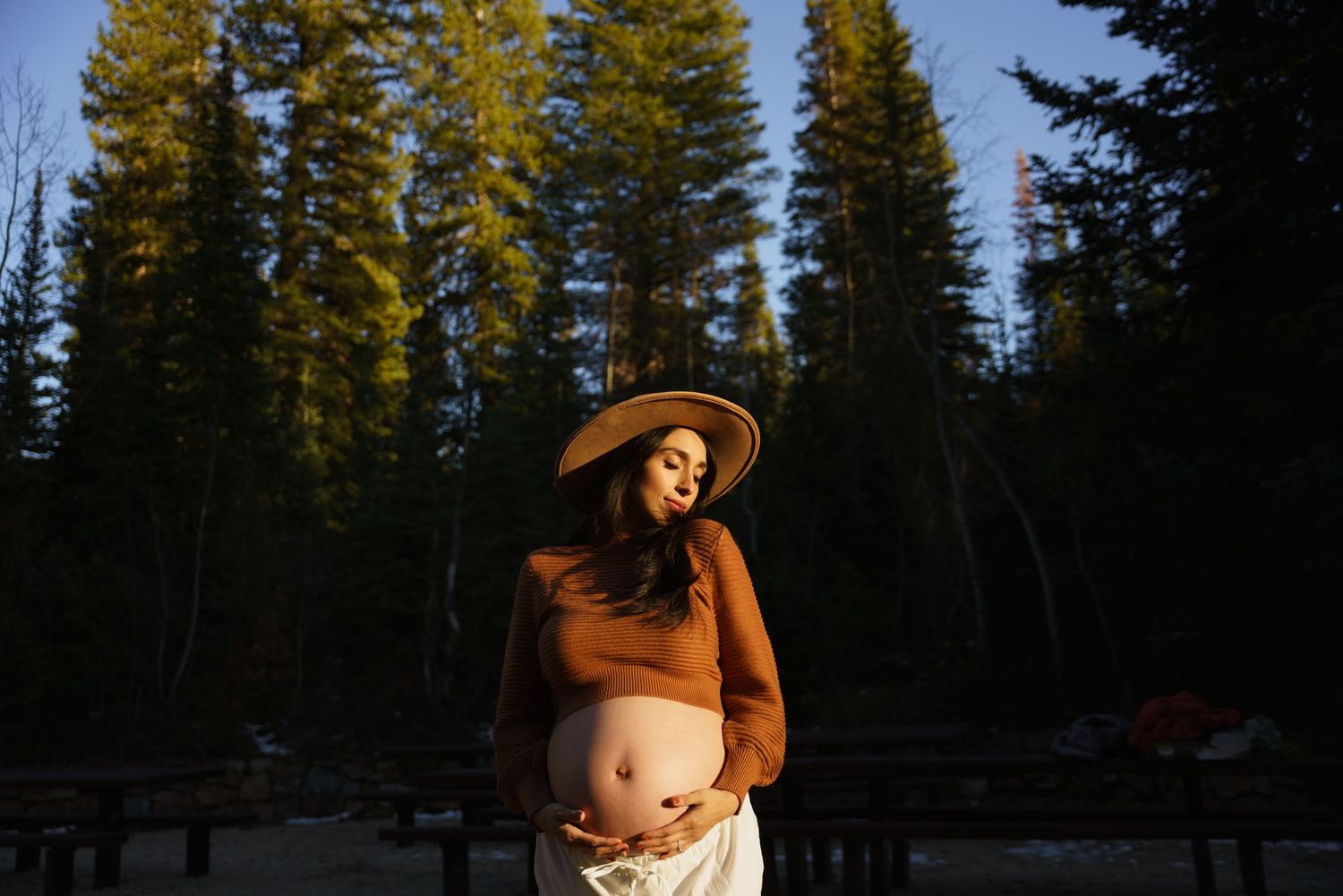 A pregnant woman in a brown top and wide-brim hat cradles her belly in a forest at sunset.