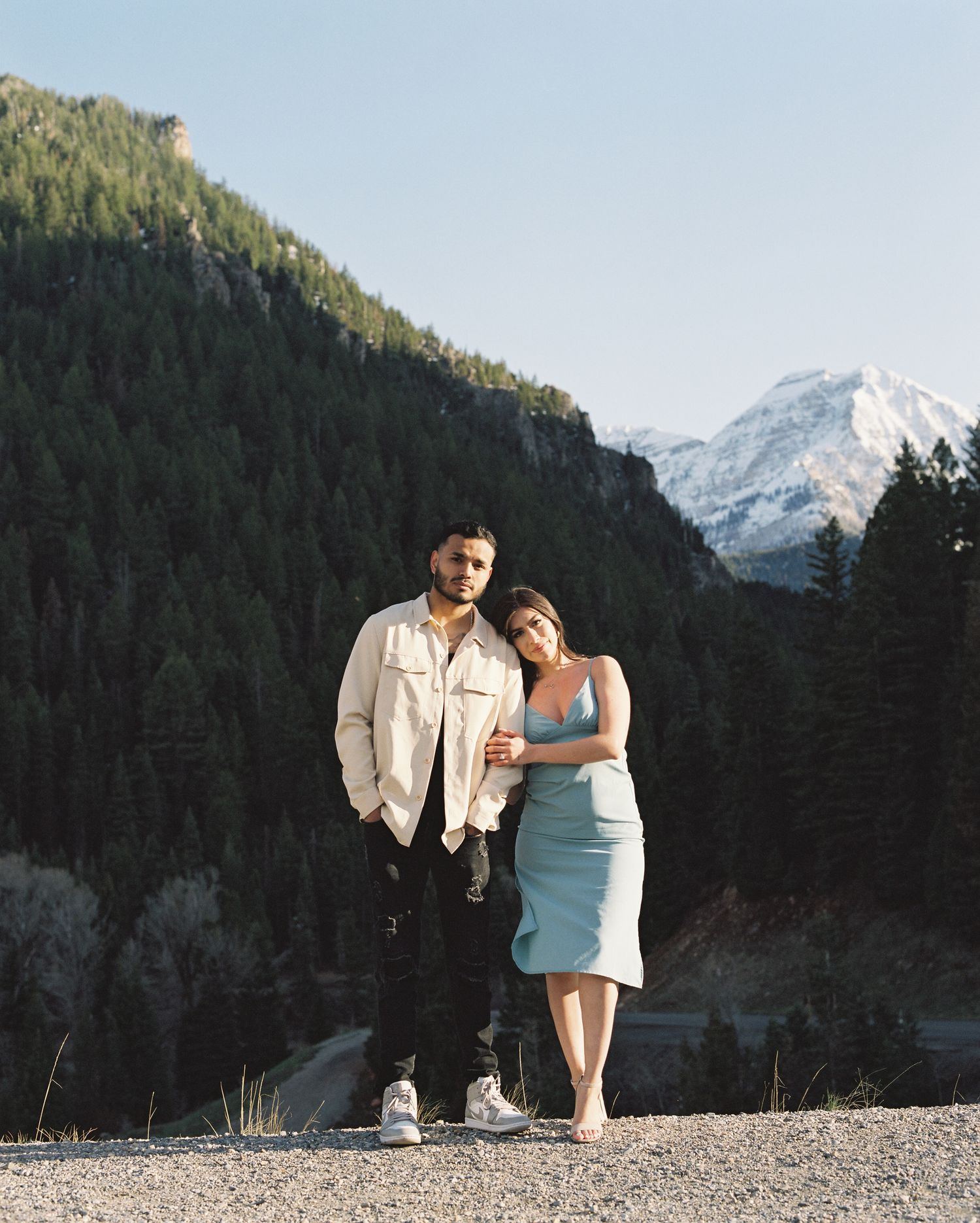 A couple poses together on a scenic mountain overlook surrounded by tall pine trees and snowy peaks.