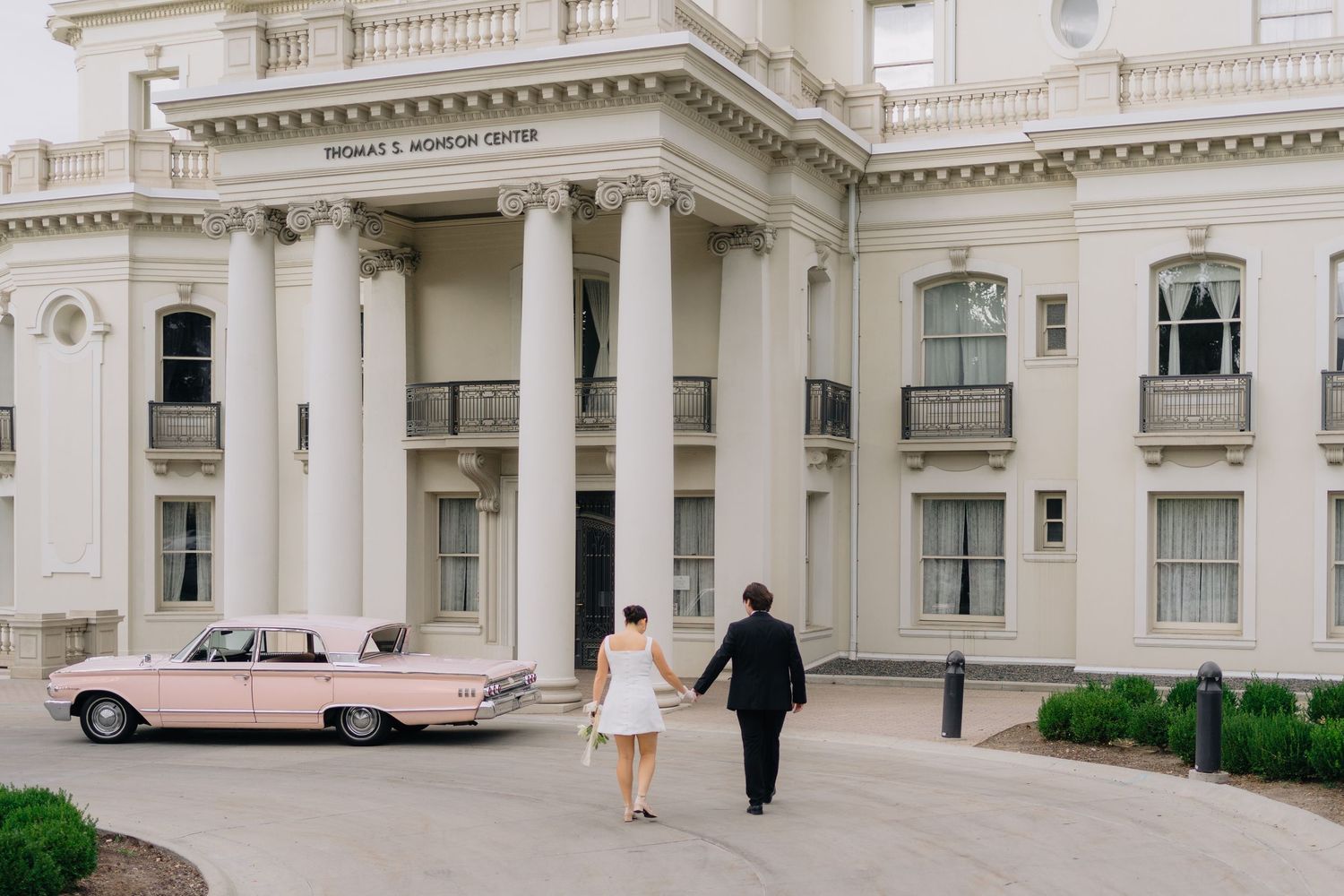 A bride and groom walk hand-in-hand toward a grand white columned building with a pink vintage car.