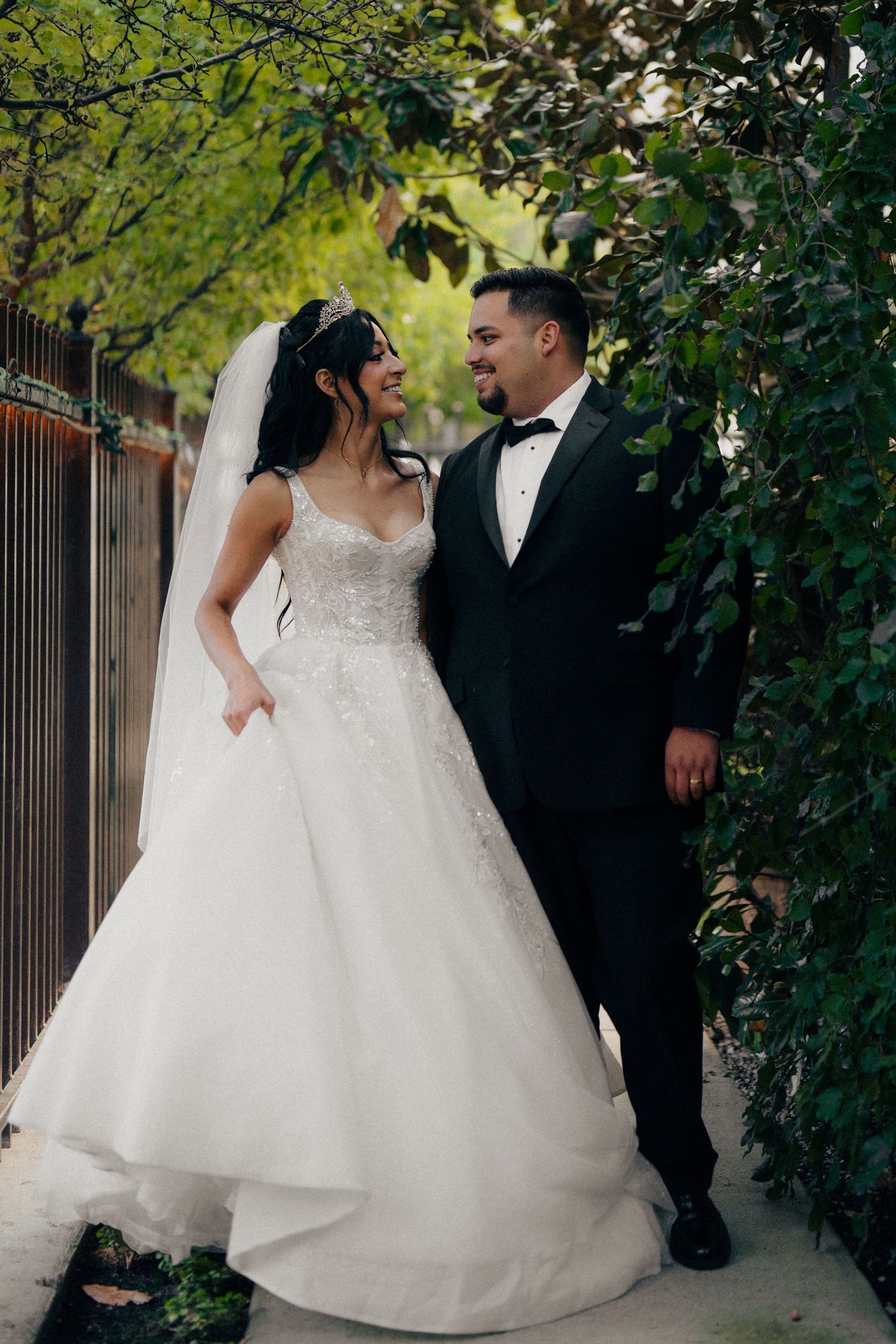 Bride in beaded ballgown and groom in black tuxedo walking under a green leafy garden arch.