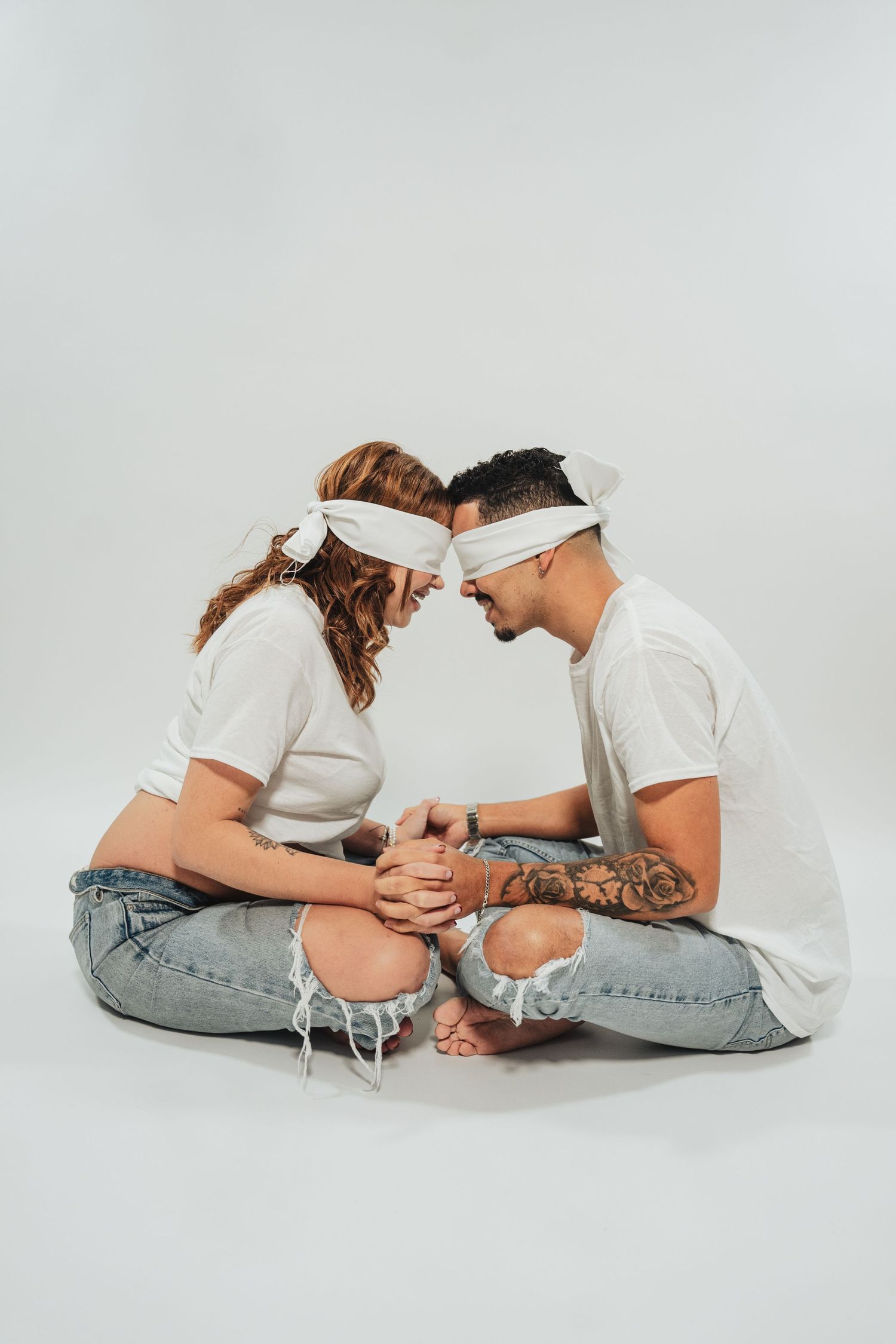 Blindfolded couple in white shirts and jeans sitting face to face holding hands on white background.