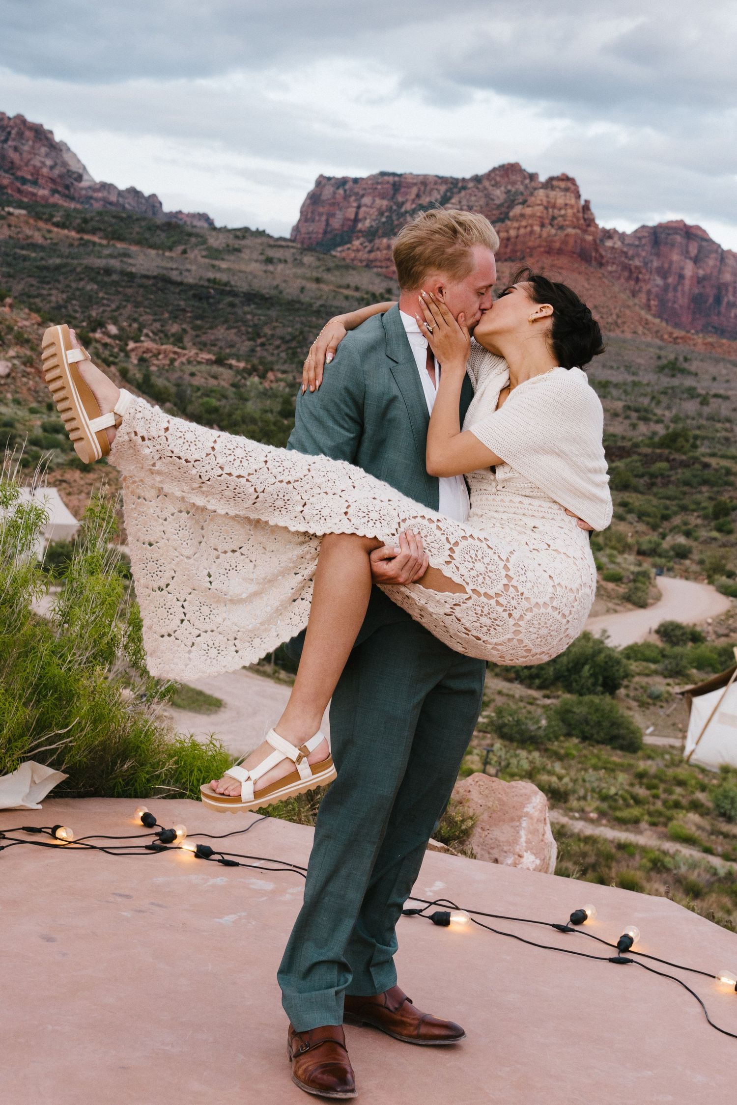 Groom lifts bride in white lace dress, sharing a kiss at a scenic Sedona red rock overlook with string lights.