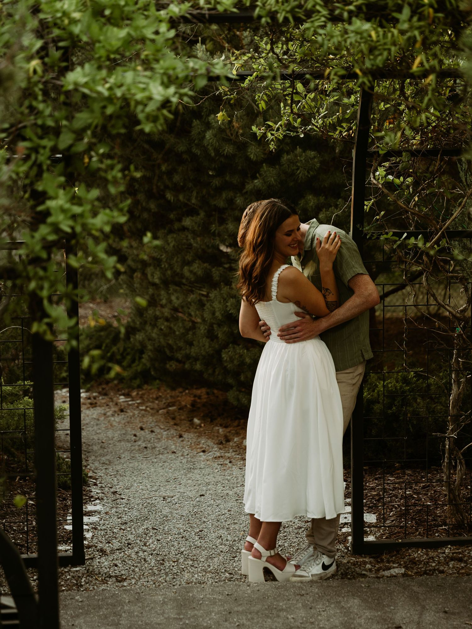 Couple sharing a romantic kiss in a lush garden doorway, woman in white midi dress and wedge sneakers.