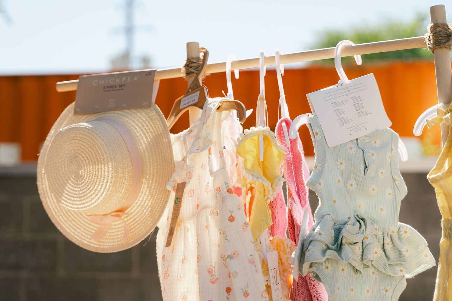 Colorful children's dresses and a straw hat hanging on a rack outdoors in sunlight.