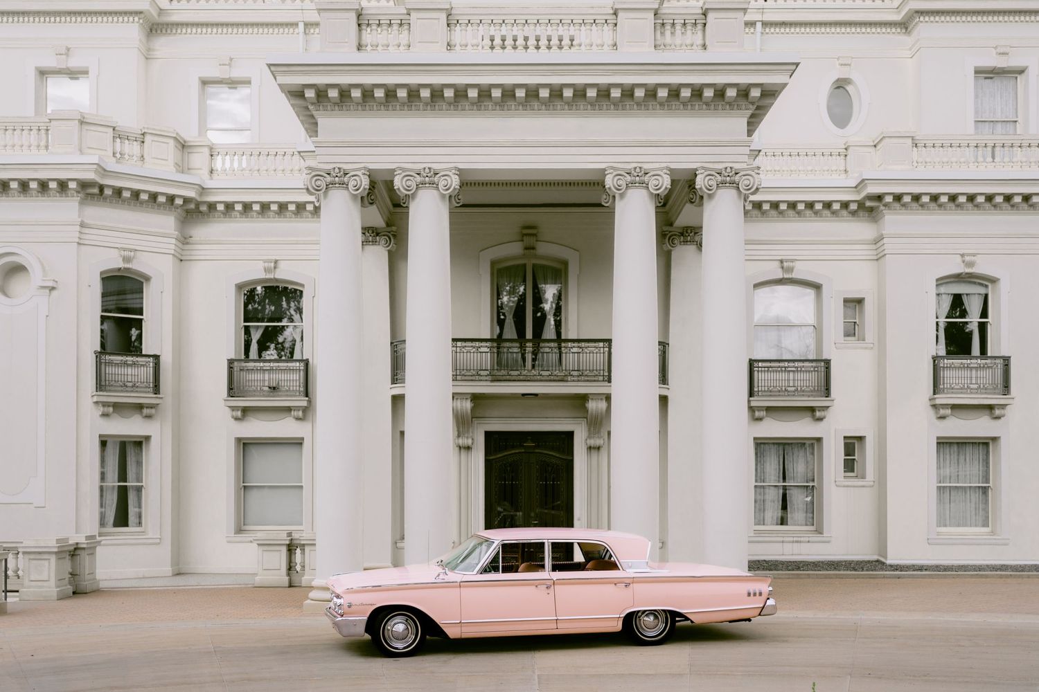 A vintage pink classic car parked in front of an elegant white columned mansion.