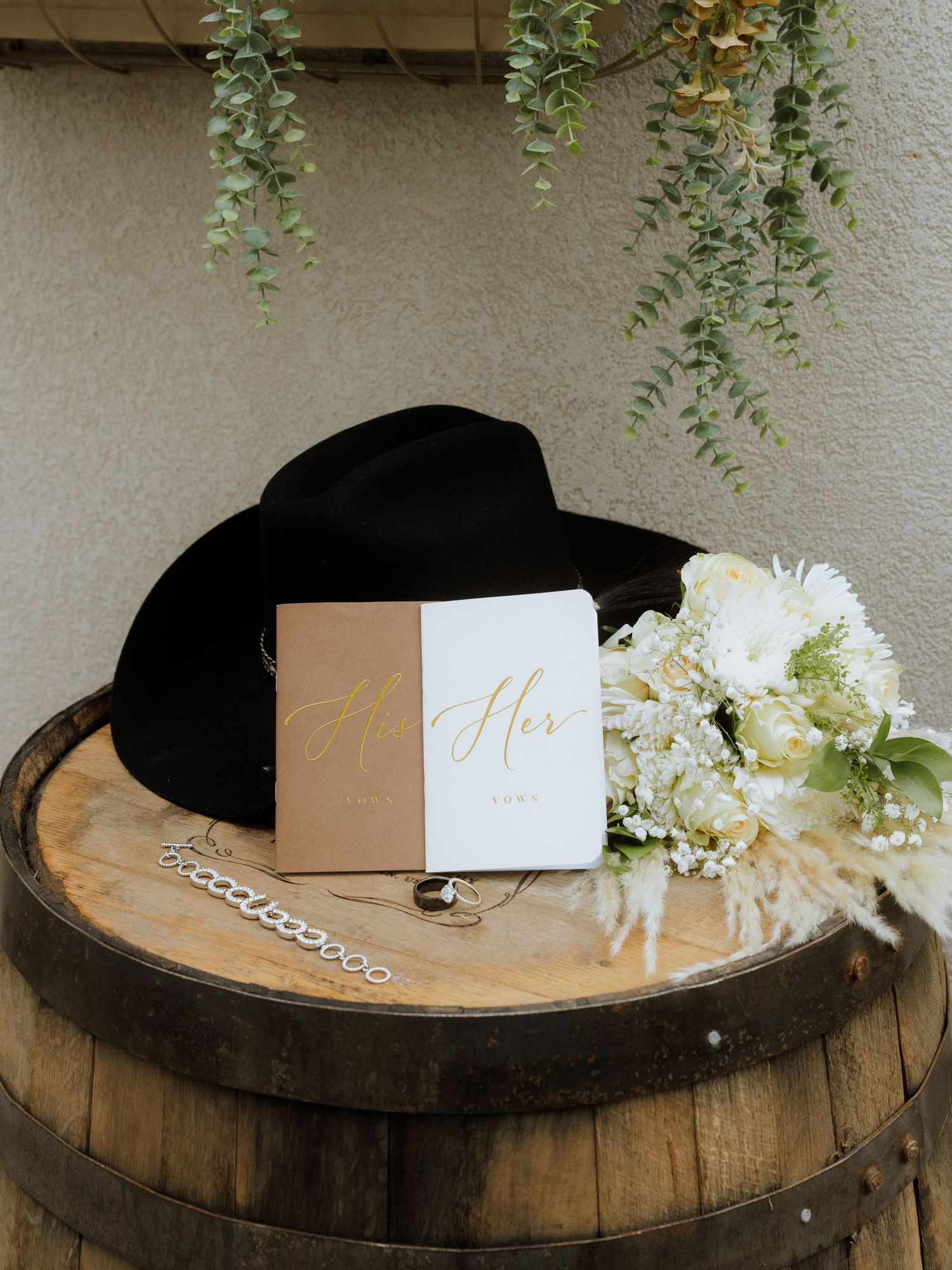 Black cowboy hat, wedding vow books, white floral bouquet, bracelet on rustic wooden barrel.