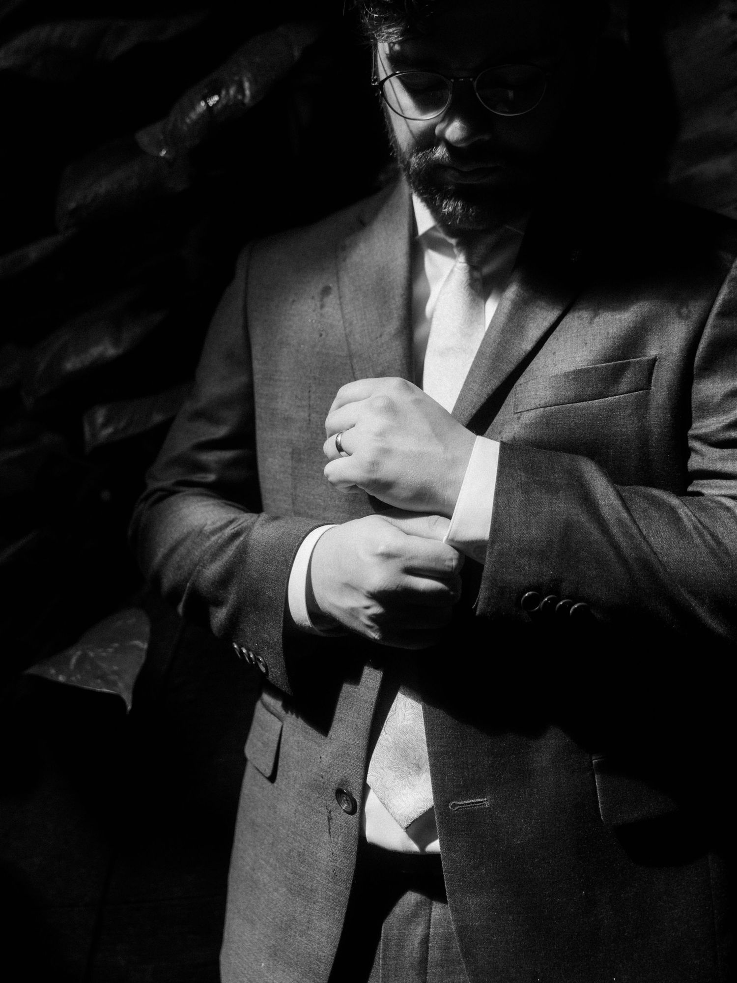 Black and white photo of a bearded man in a suit adjusting his cufflinks, wearing sunglasses.