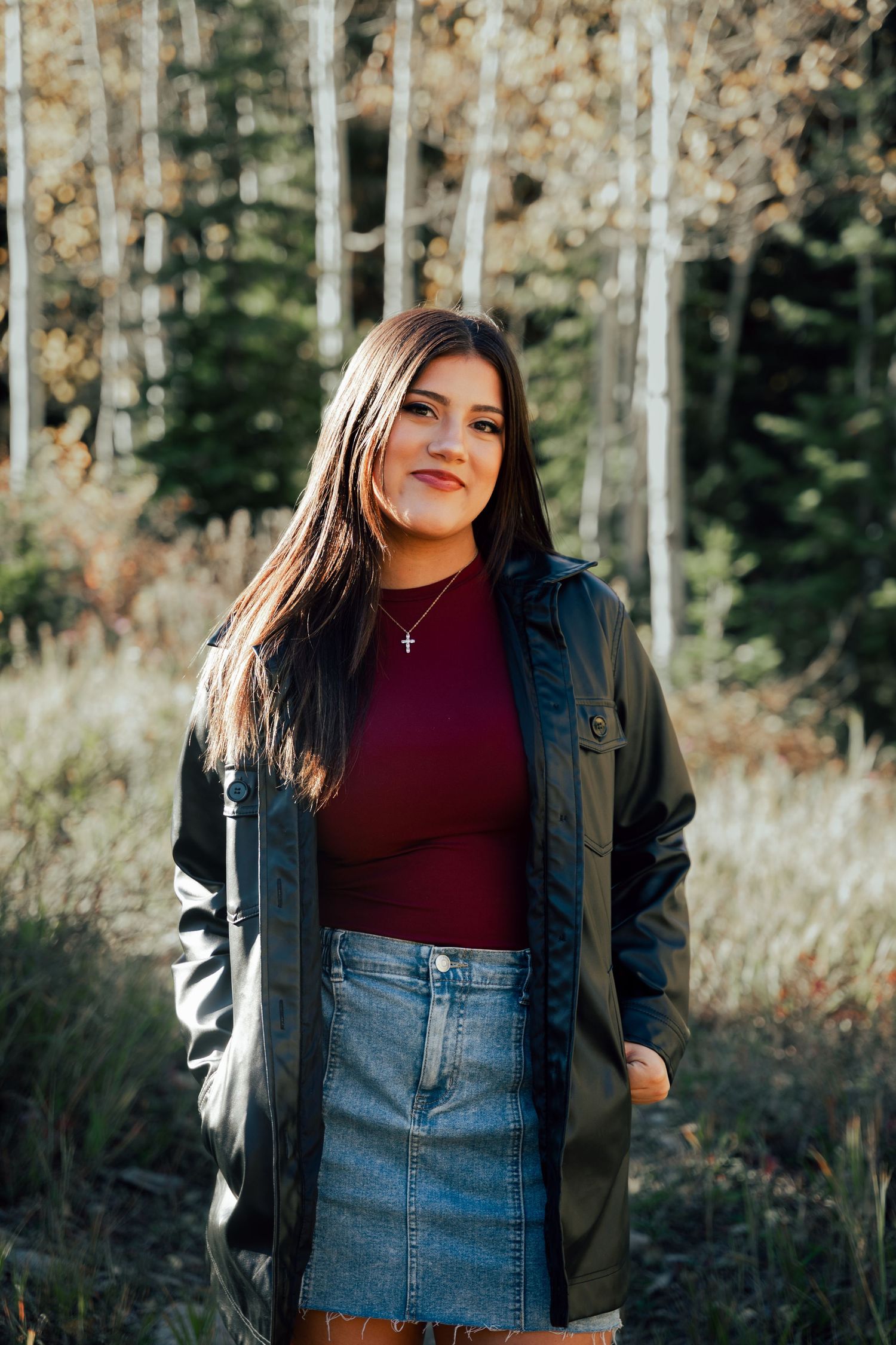 Smiling young woman in burgundy top, denim skirt and olive jacket posing in autumn forest.