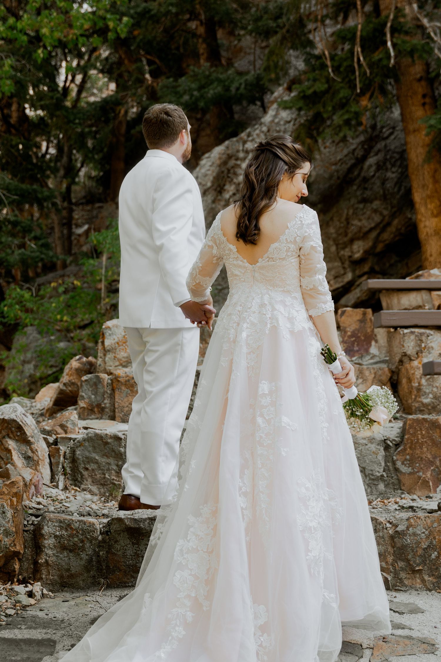 A bride in a lace gown and groom in white suit hold hands in a rocky forest setting.