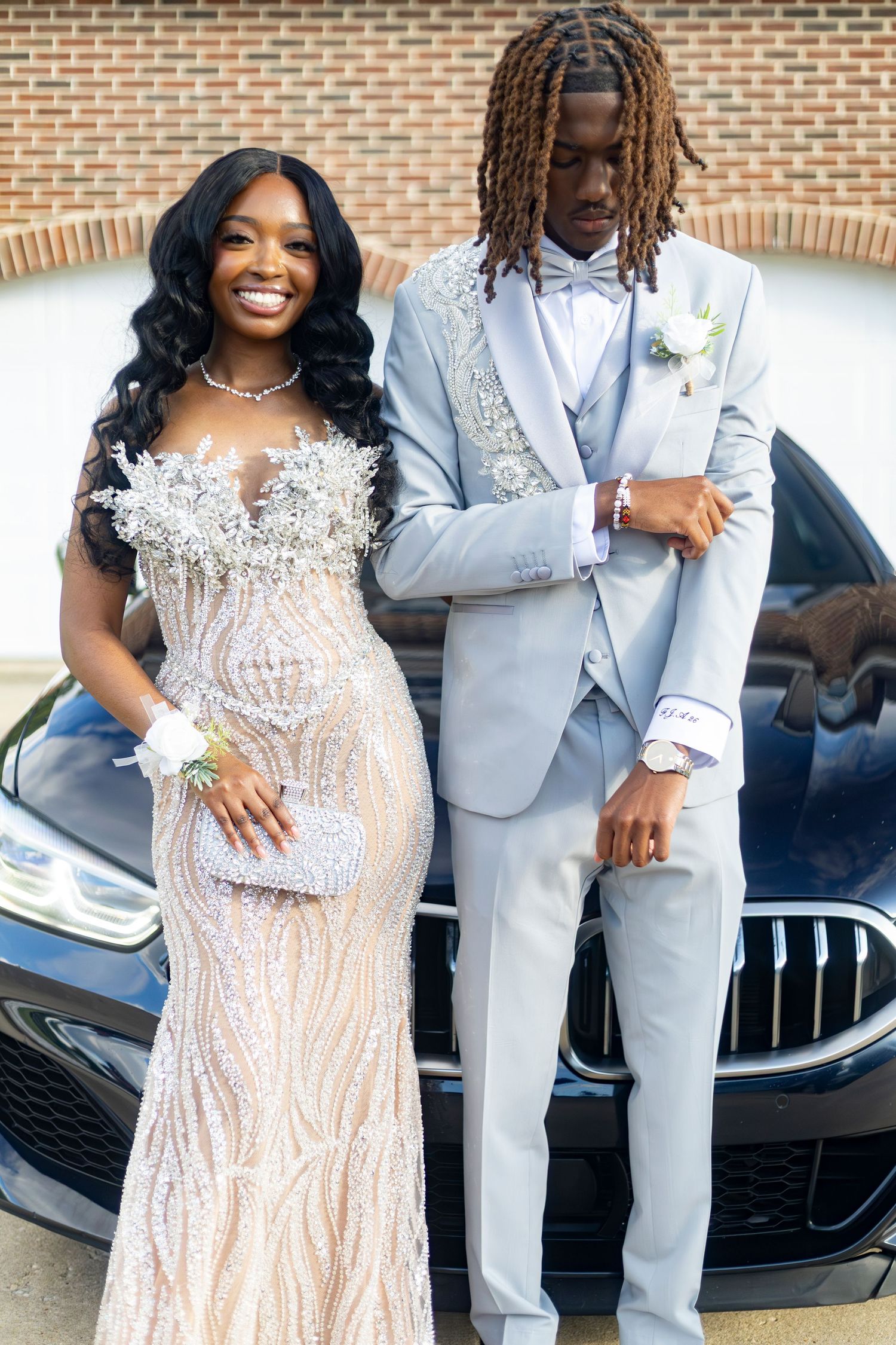 A young couple posing in elegant prom attire in front of a luxury car.