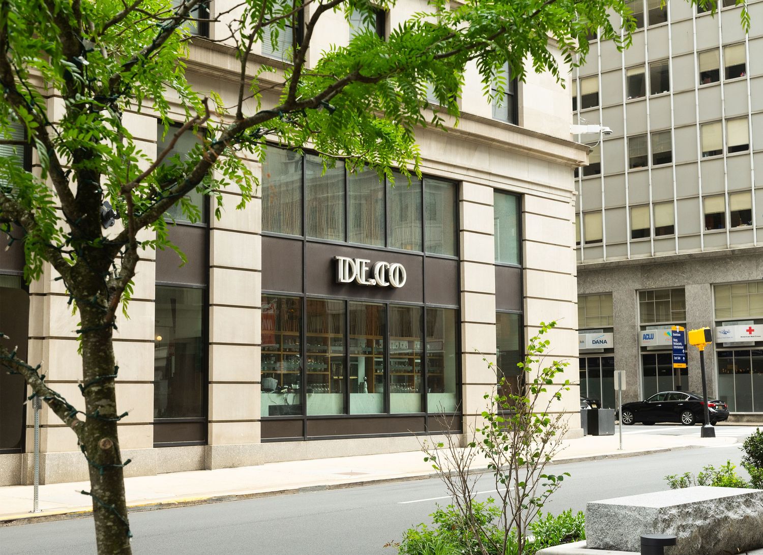 An exterior view of a city building storefront with DE.CO signage, framed by green trees and captured by a professional landscape photograph
